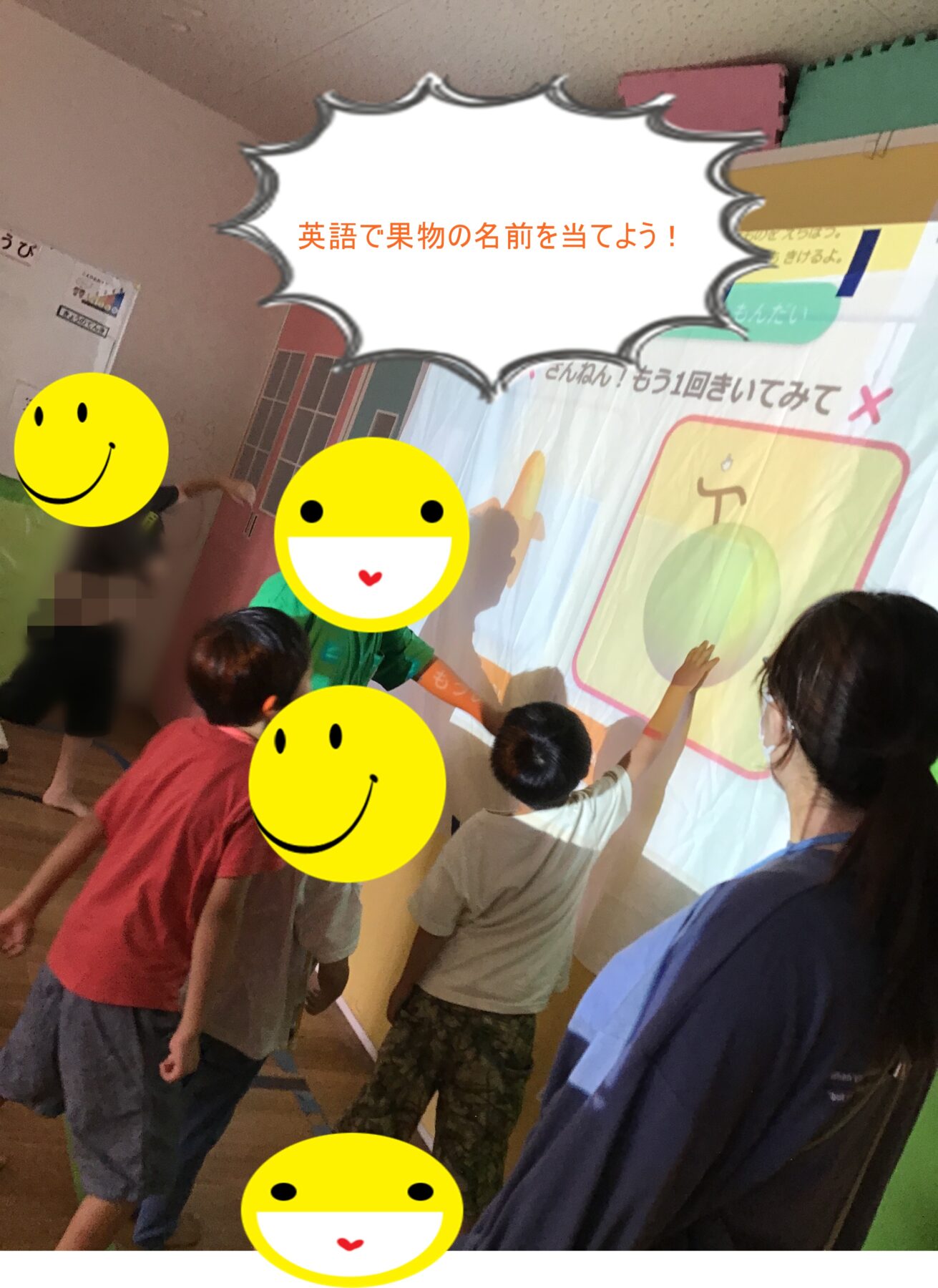Children in a classroom line up at a wall display about fruit names in English, with a large speech bubble asking to guess the fruit names.