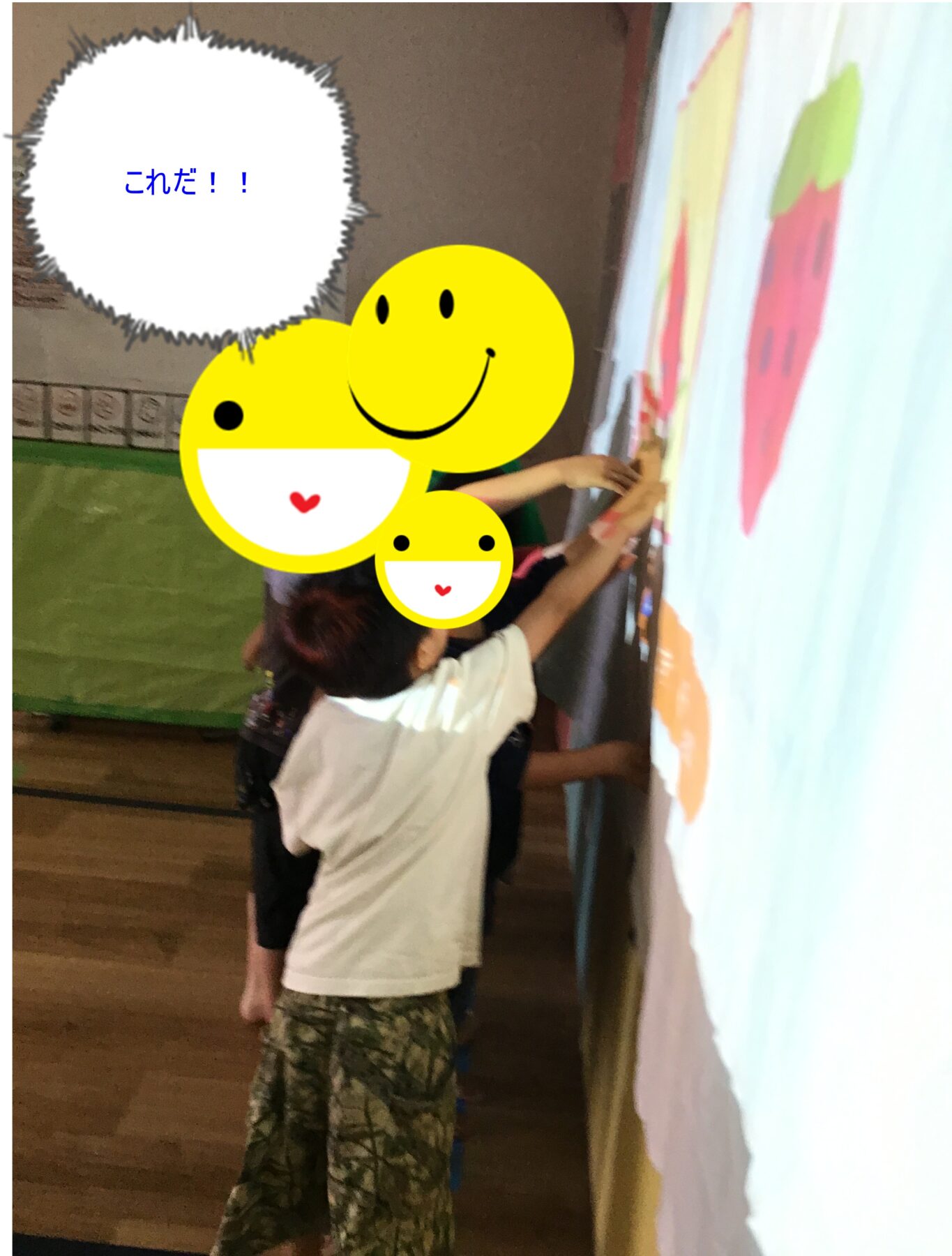 Children in a gym or classroom pasting large yellow smiley stickers onto a wall mural with a speech bubble reading これだ!!