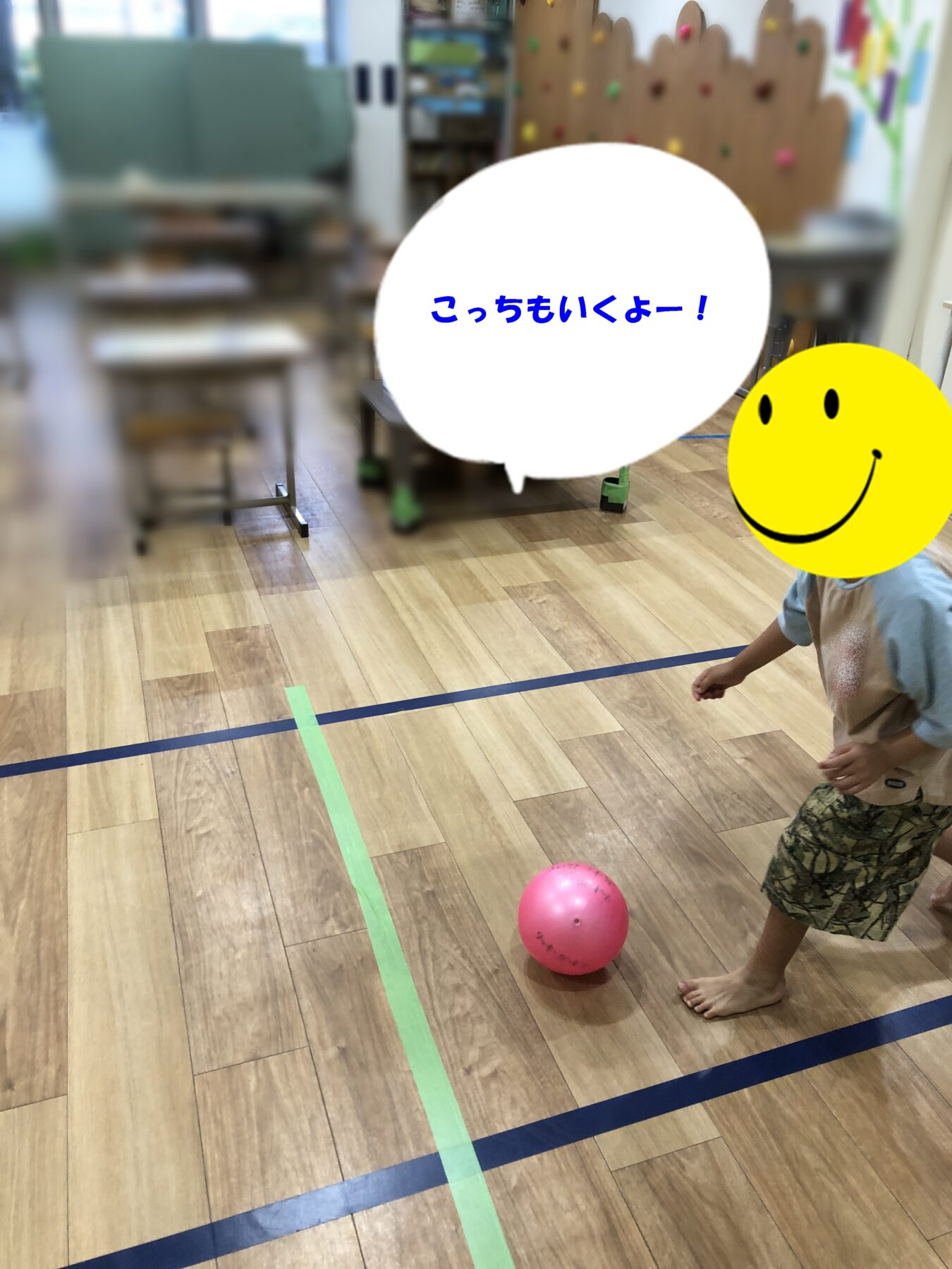 Child playing with a pink ball on a wooden gym floor, marked by green tape lines; blurred playroom background with a large white speech bubble and a bright yellow smiley graphic partially covering the scene.