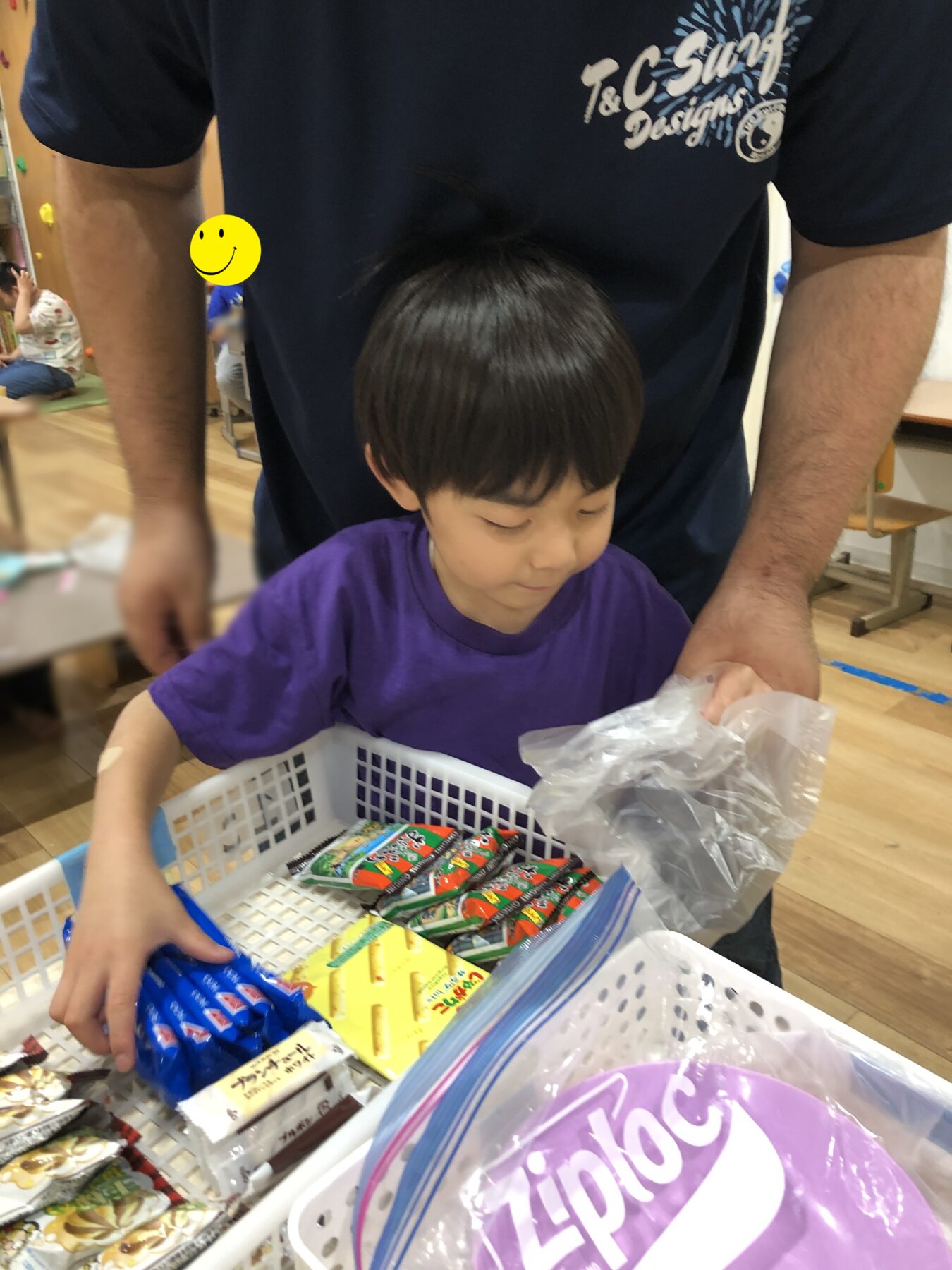 Young child in a purple shirt reaches into a white basket filled with colorful snack bars, with an adult standing behind.