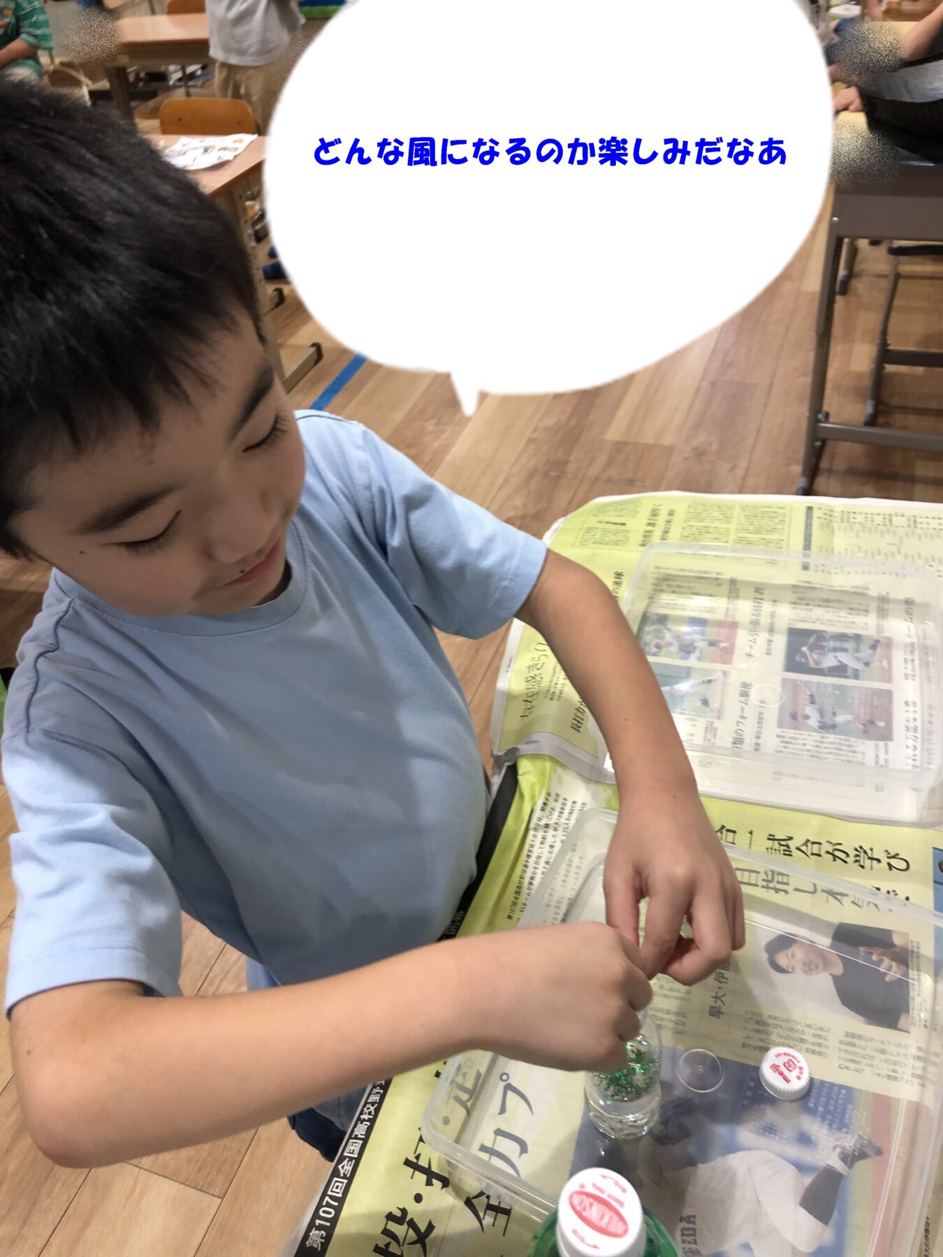 Young boy in a light blue shirt sits at a table with a newspaper-covered surface, threading beads from small containers in a classroom.