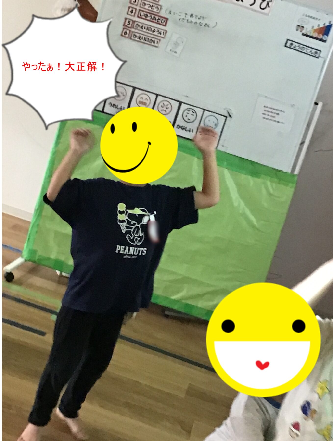 Child celebrating with arms raised in a school gym, wearing a dark Peanuts shirt; a white speech bubble with red Japanese text 'やったぁ!大正解!' and a large yellow smiley sticker obscuring part of the scene.