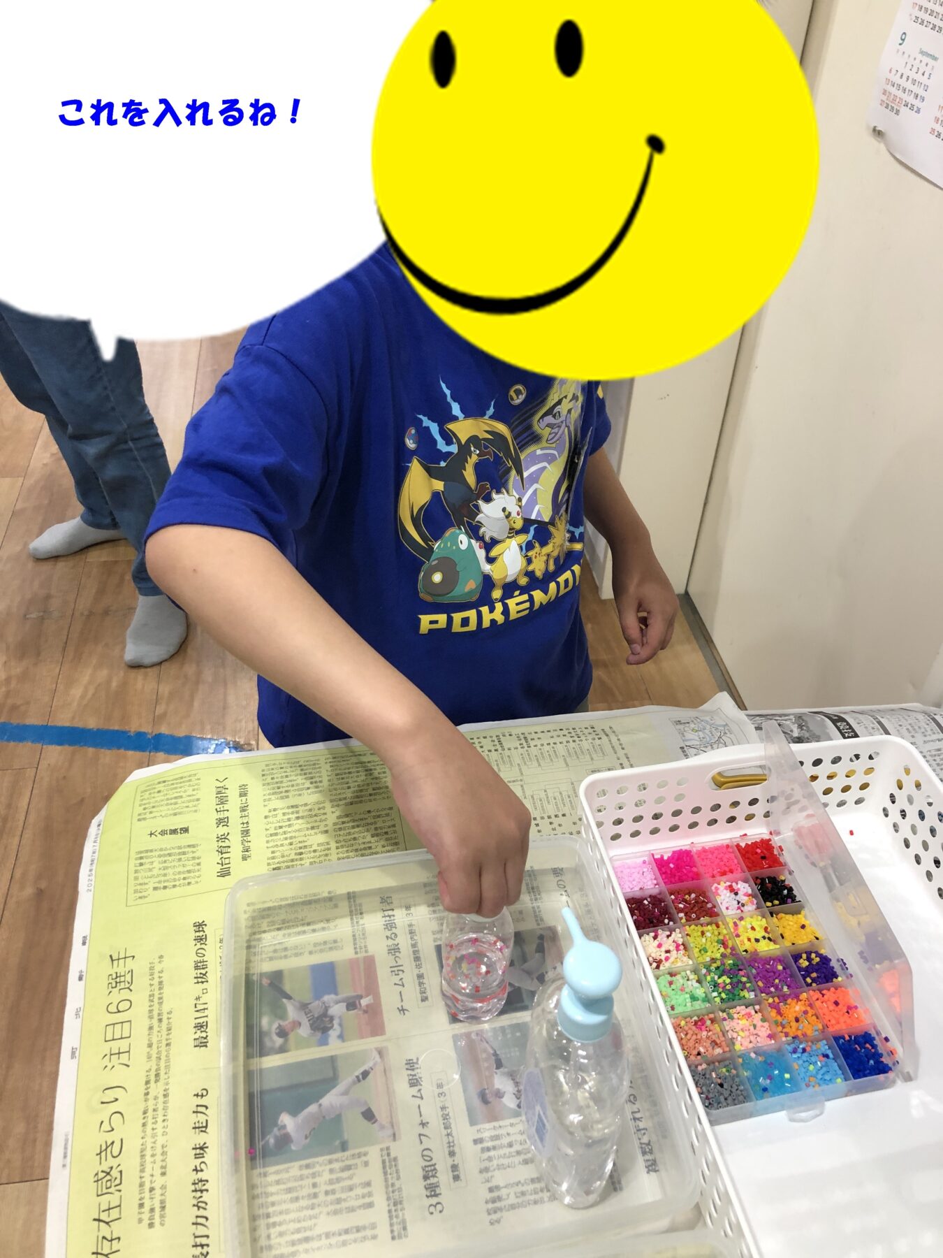 Child in a blue Pokémon shirt sorts multicolored bead beads from a plastic organizer onto a newspaper-covered table with a small cup nearby