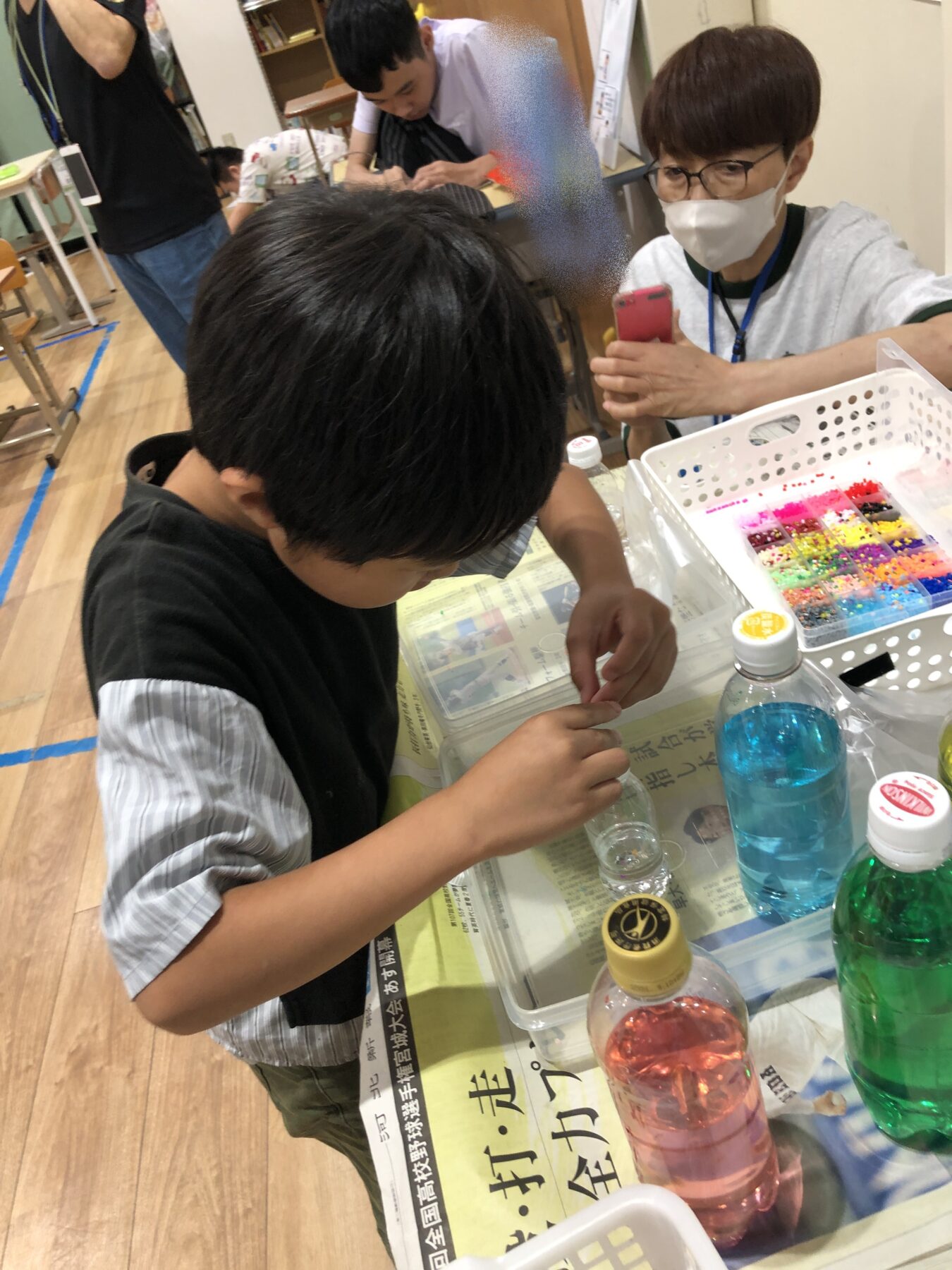 Child arranging colorful beads at a craft table with a supervising adult nearby, beads organized in a tray and bottles on the table behind him.