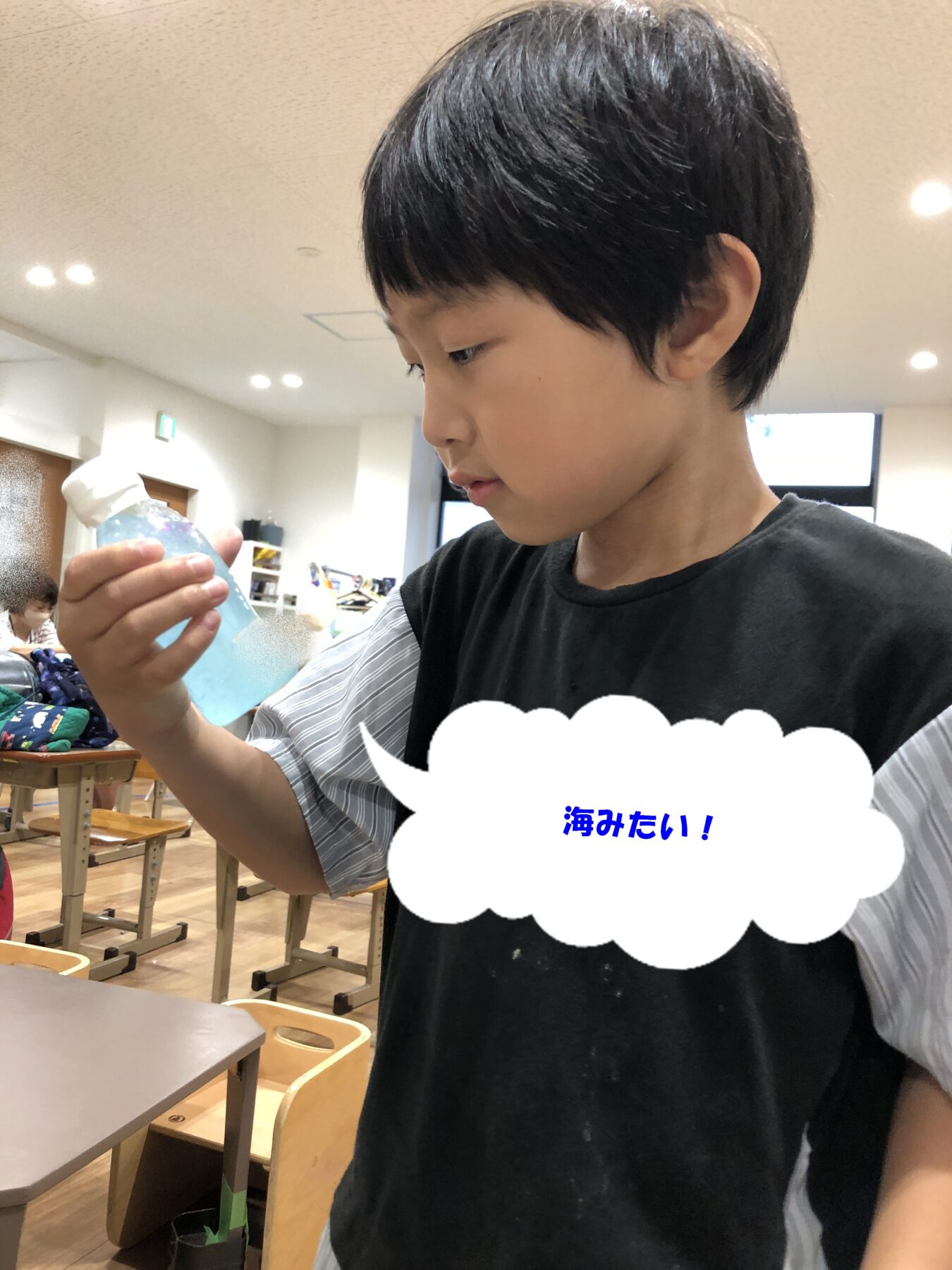 Boy in a black shirt holds a blue plastic bottle up, looking at it in a bright classroom with desks and chairs in the background.