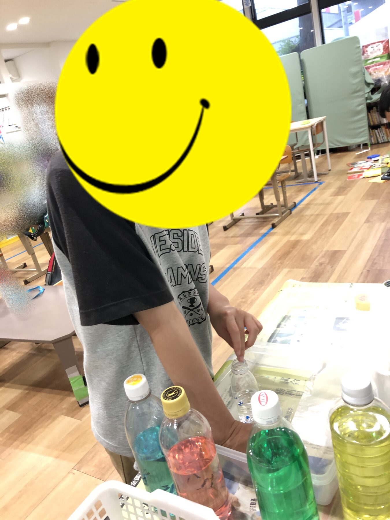 Child standing at a table with plastic bottles and colorful drinks in a classroom or library; a large yellow smiley graphic covers their face.
