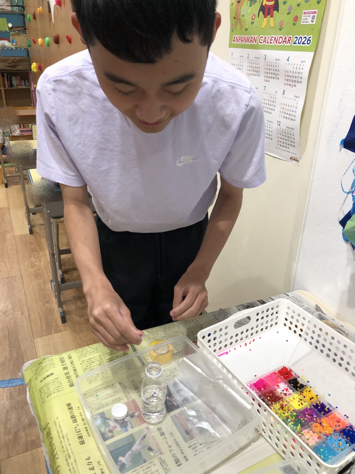 Student standing at a table arranging small colorful beads in a clear organizer, with a newspaper-covered workspace nearby in a classroom setting.