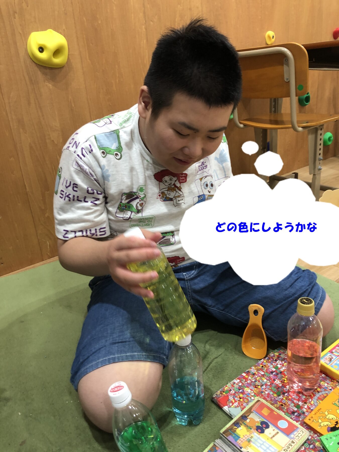 Young boy sitting on the floor, holding a yellow bottle, surrounded by colorful books and bottles; a climbing wall in the background.
