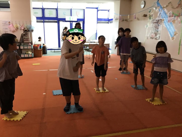 Group of children standing on colored foam tiles arranged in rows inside a bright classroom, ready for a game or activity. One child in the foreground has a cartoon sticker over their face for privacy.