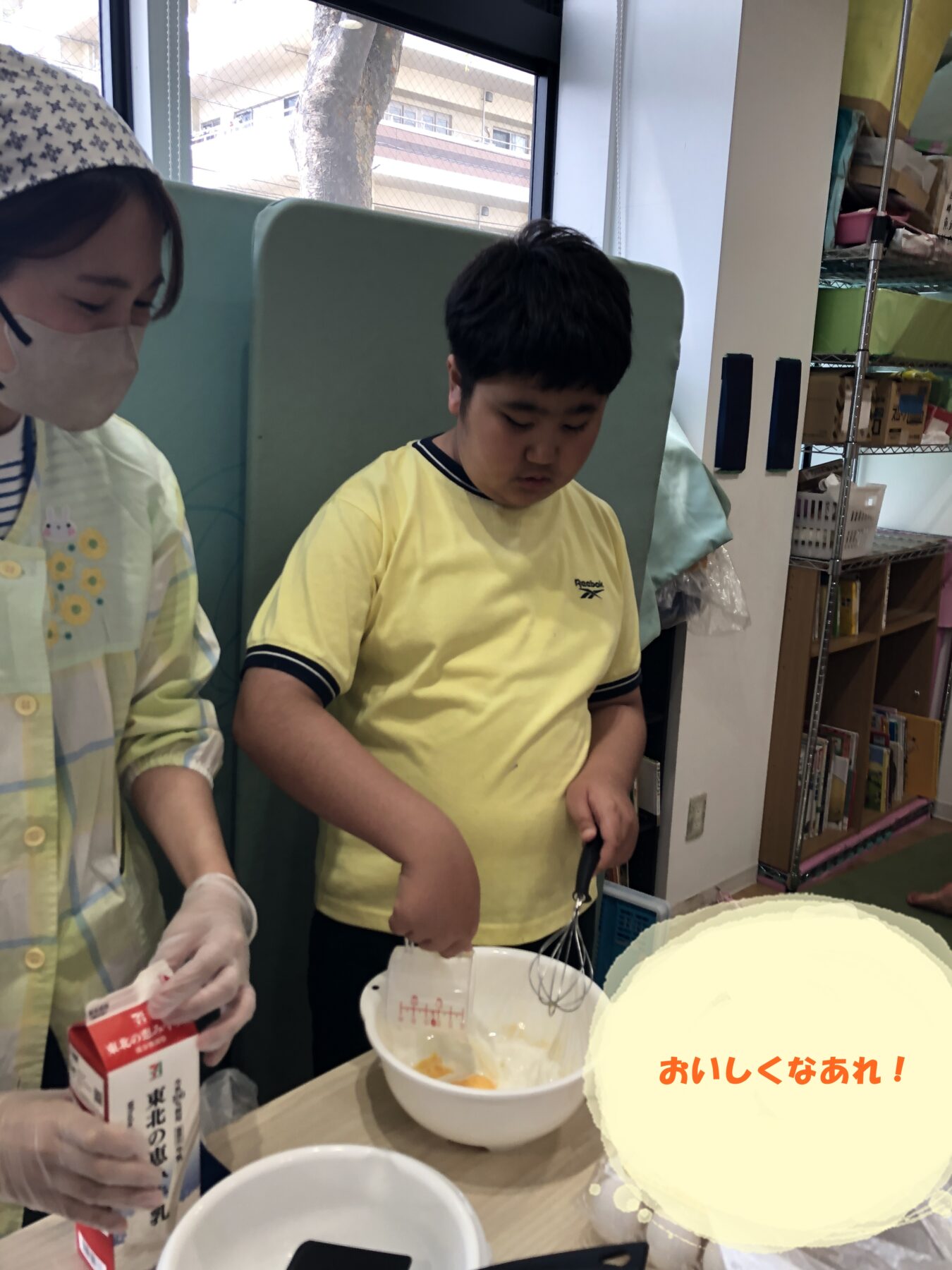 Boy in a yellow shirt uses a handheld mixer to whisk batter in a bowl, assisted by an adult wearing a mask in a bright room with shelves in the background.