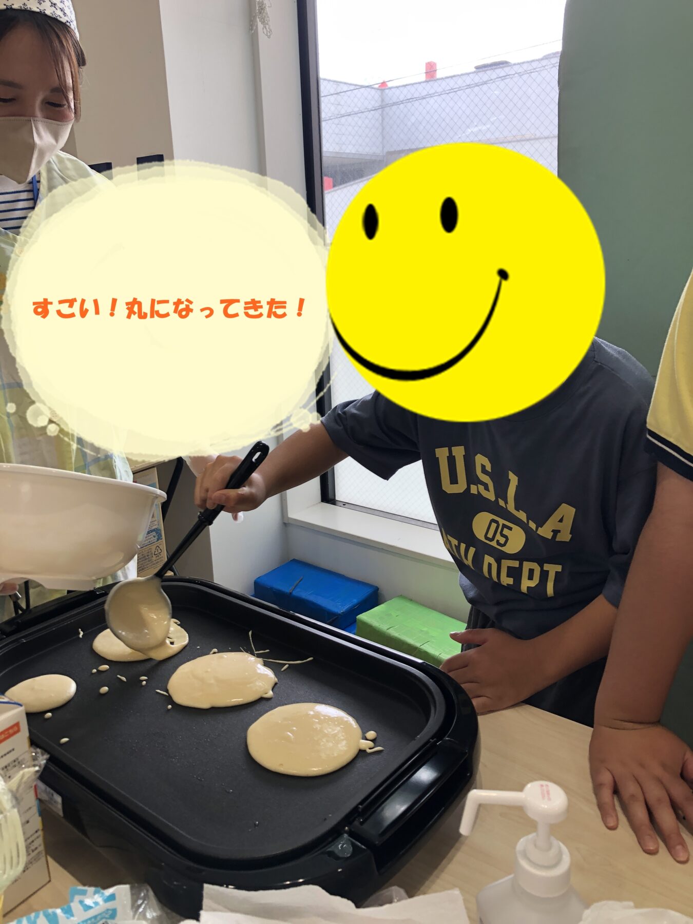 Child wearing a mask watches as pancake batter cooks on an electric griddle, forming round circles.