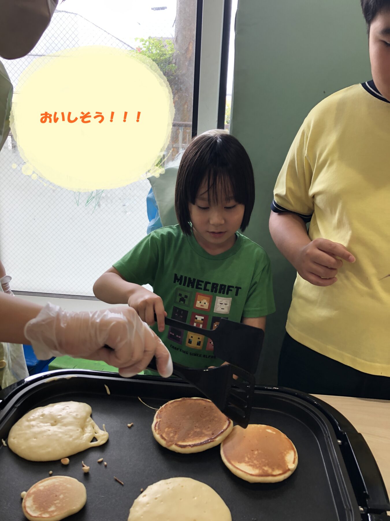 Children cooking pancakes on a griddle; a kid in a green Minecraft shirt flips batter with a spatula while others help, with a large yellow speech bubble showing 'おいしそう!!!' in the background.