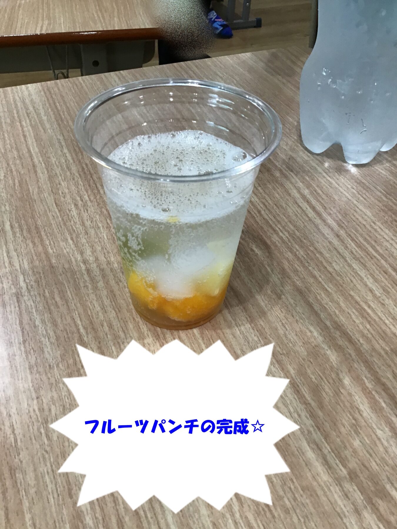 Clear plastic cup of fruit punch with orange fruit pieces and bubbles on a wooden table, foreground focus on drink.