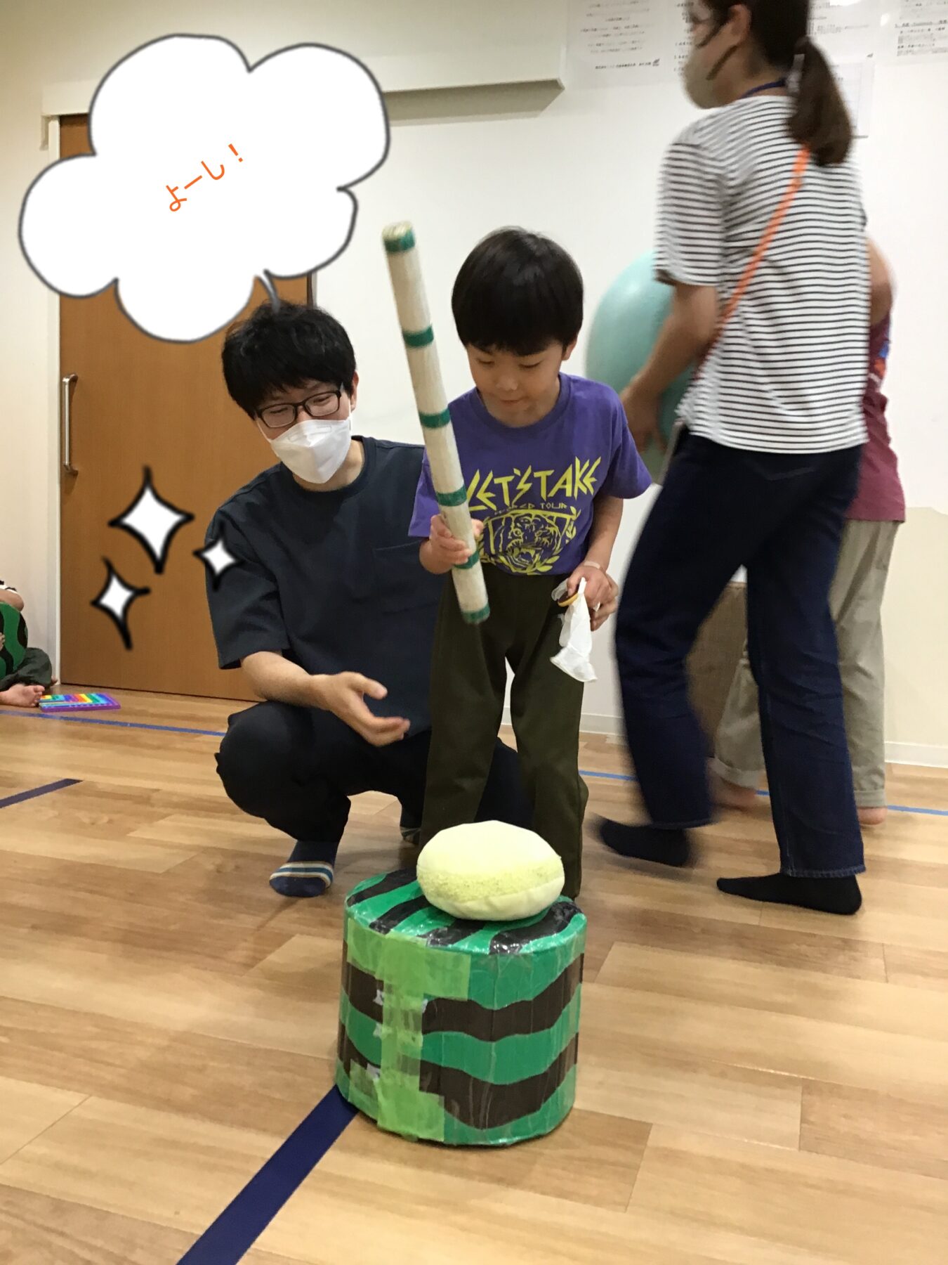 Child in a purple shirt holds a long striped baton, aiming at a foam target atop a taped green-and-brown stacked cylinder, while an adult in a mask crouches nearby; speech bubble reads よーし！