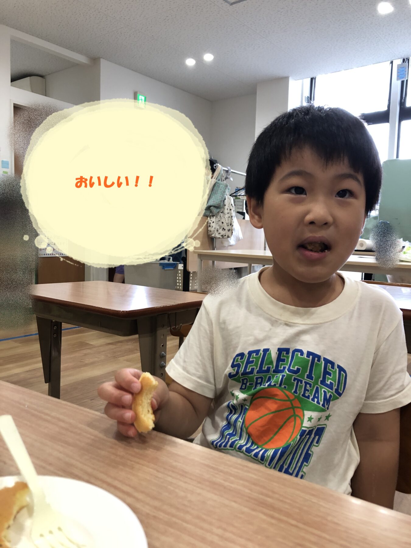 A young boy sits at a cafeteria table, biting into a snack while wearing a white t‑shirt with a basketball graphic, in a bright room. plate with food is visible in the foreground and other tables are in the background.