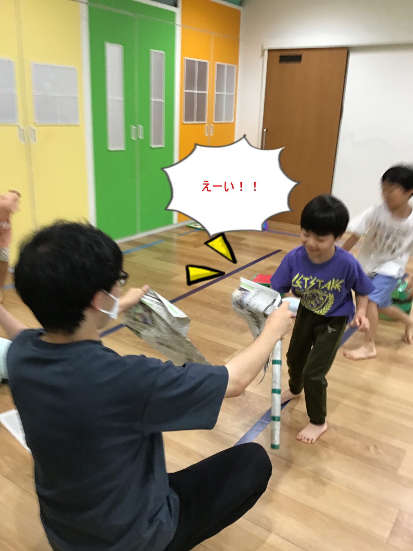 Boy in a purple shirt smiles as an adult kneels and hands him a newspaper during a playful indoor activity, with yellow, green, and orange lockers in the background and a comic-style speech bubble saying 'えーい!!'