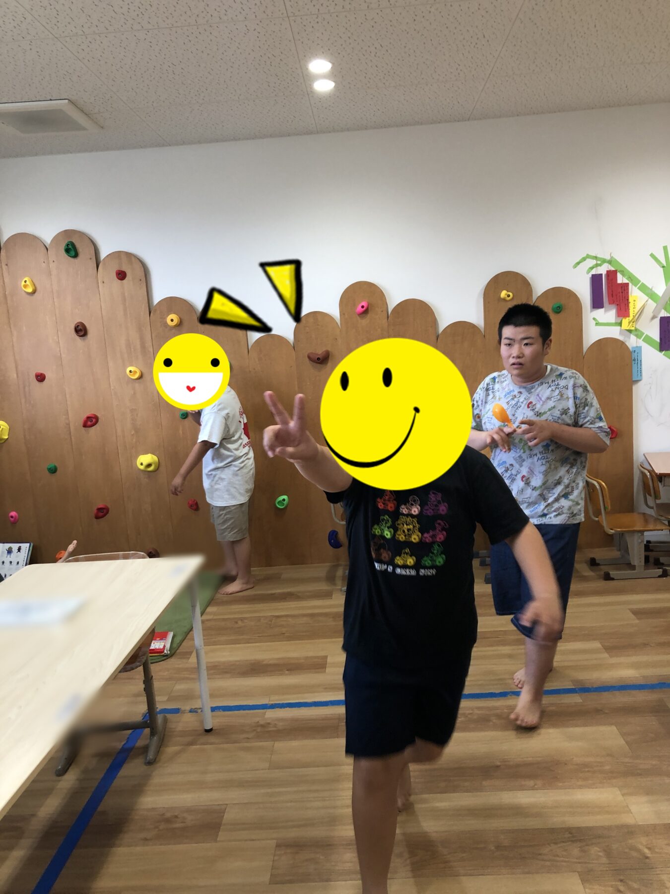 Indoor play area with a wooden climbing wall and colorful handholds; children playing while a person in the foreground makes a peace sign.