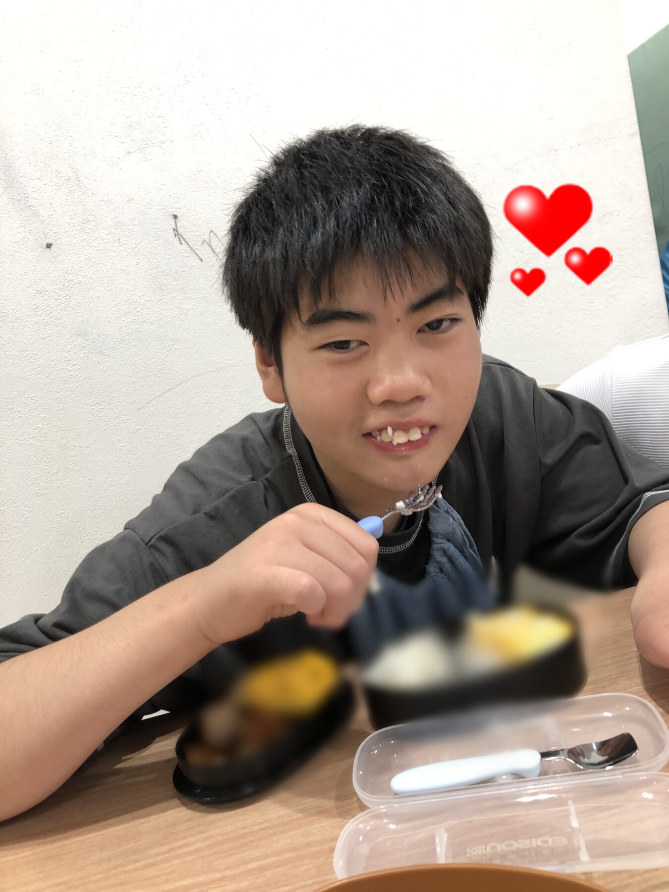 Smiling boy uses chopsticks to eat sushi, with red heart stickers in the background.