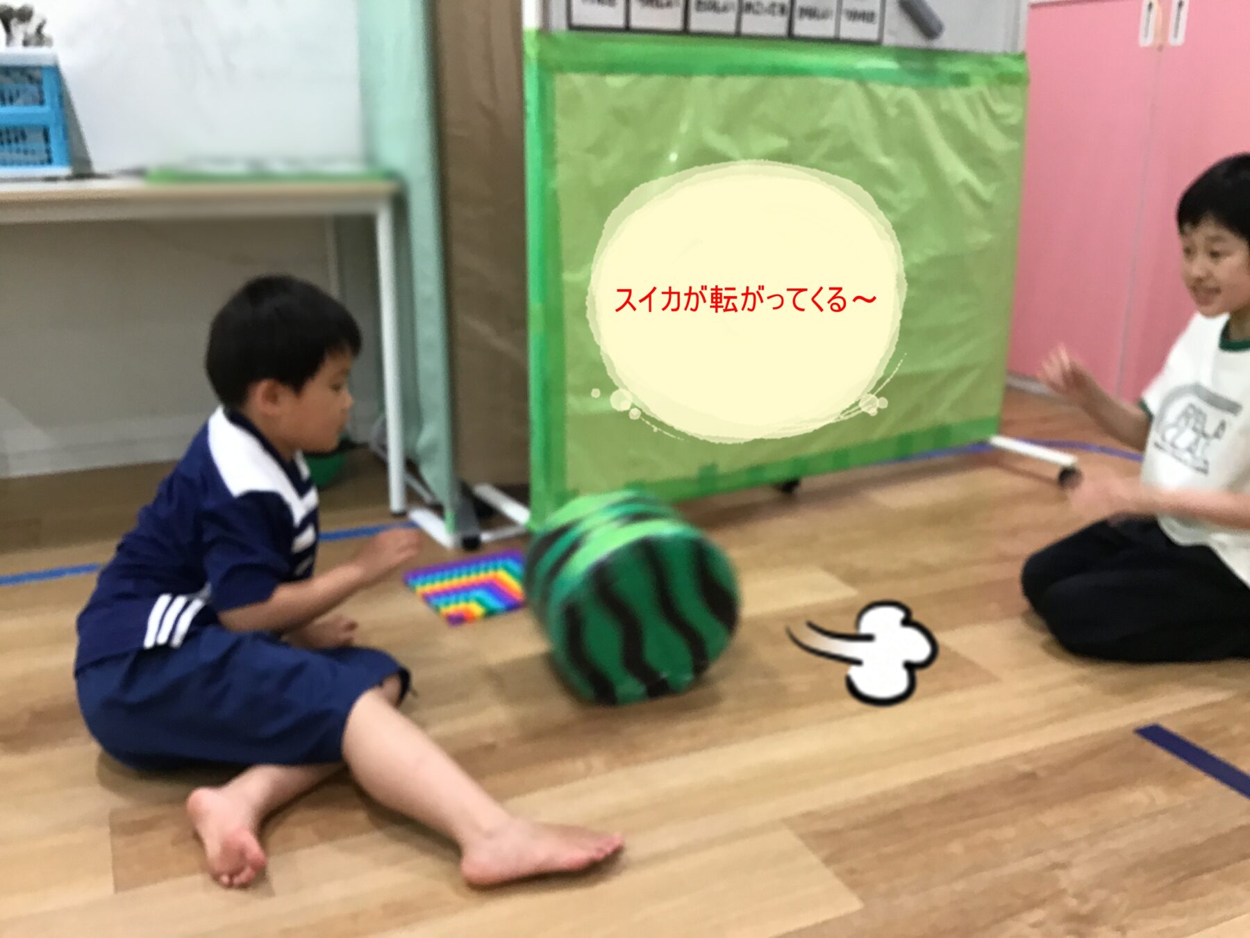 Two children sit on a wooden floor playing with a green striped ball; a pink cabinet and a green-wrapped box are in the background, with a speech bubble that reads “スイカが転がってくる〜.”