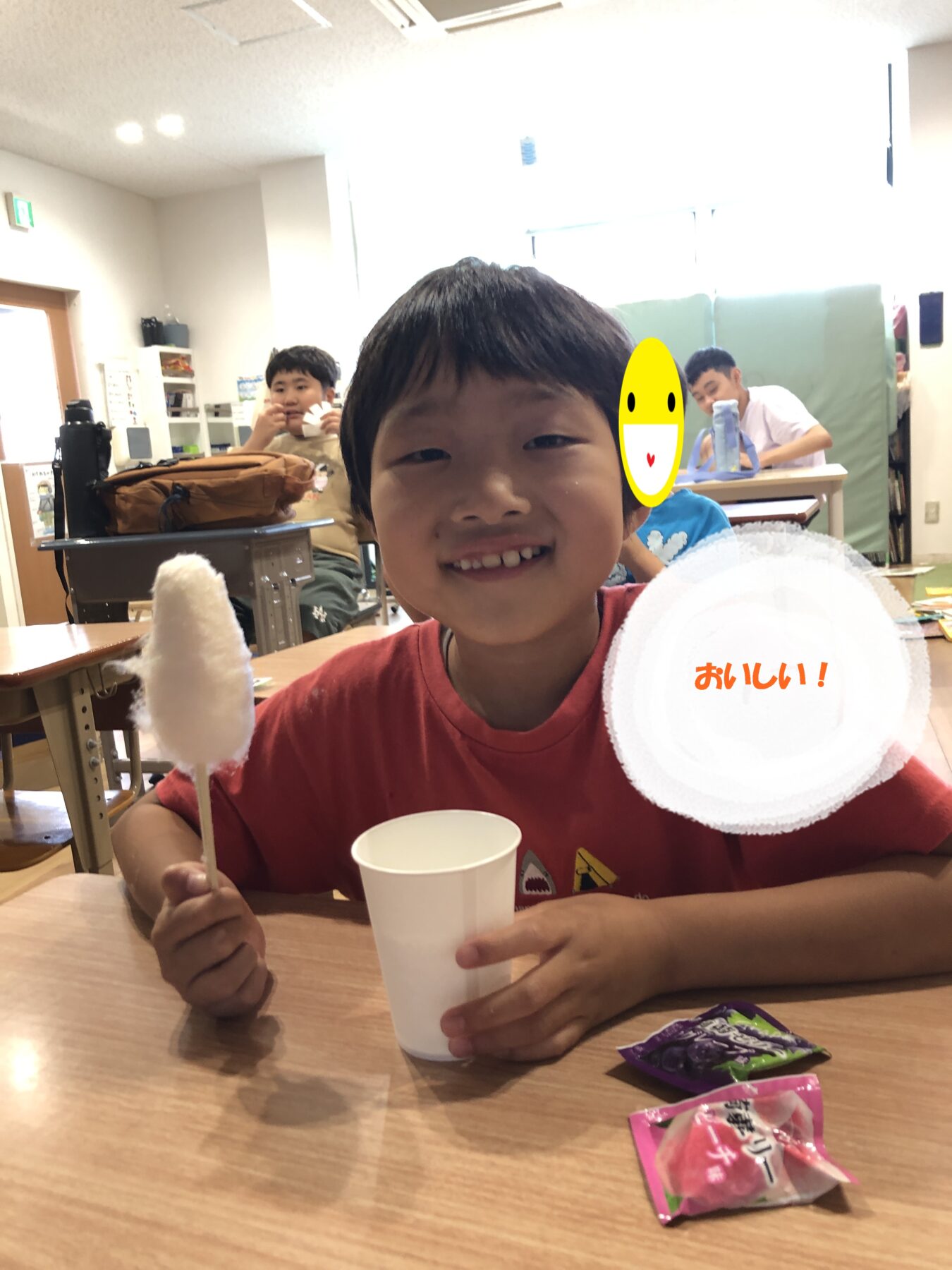 Smiling boy in a red shirt at a classroom table holding cotton candy on a stick, with a white cup; background kids and a circular overlay reading 'おいしい！'