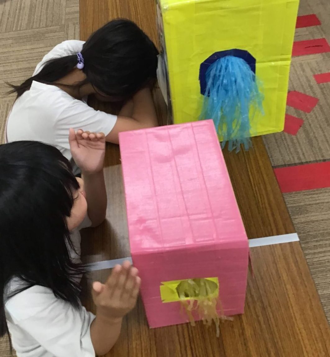 Children sitting on a carpeted floor, playing with large painted cardboard boxes for a classroom craft activity.