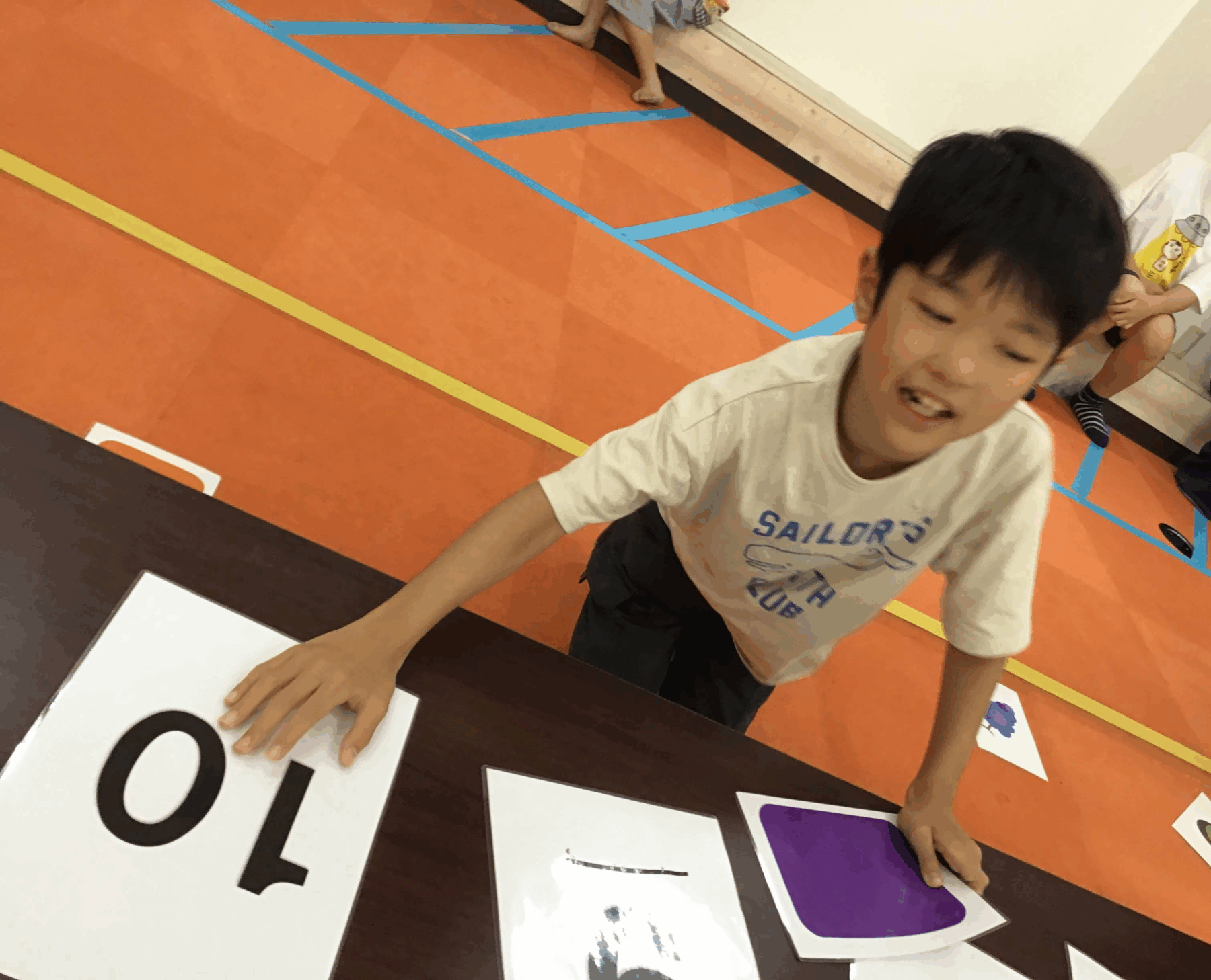 A boy leaning over a table places a large white card with bold black numbers during a classroom activity on an orange gym floor with blue tape lines in the background.
