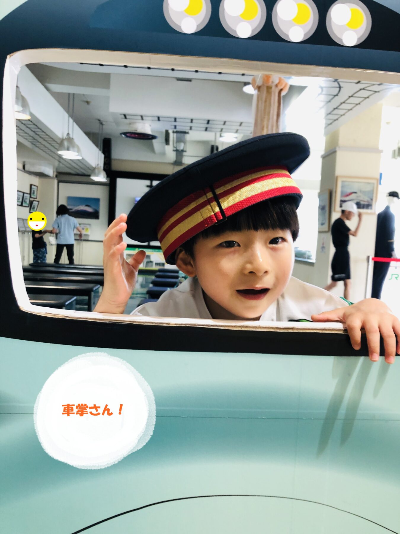 Boy wearing a black captain's hat with red-gold band peeks through a blue display window, waving his hand; round white text bubble with red Japanese characters is visible below.