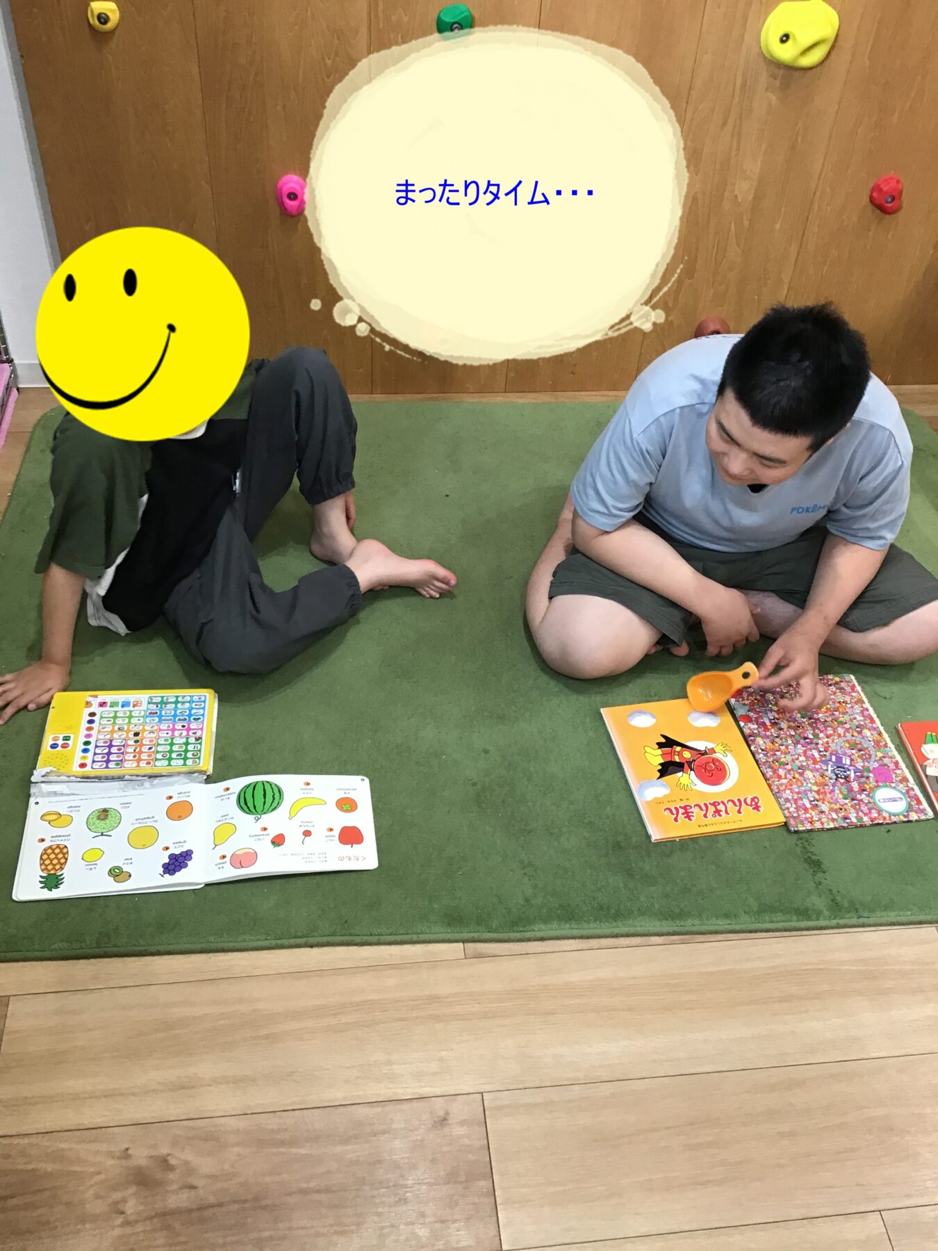 Two people sit on a green mat, surrounded by colorful children's books and puzzles; one masked by a smiley face, text in Japanese reads 'relax time' (informative, casual scene).