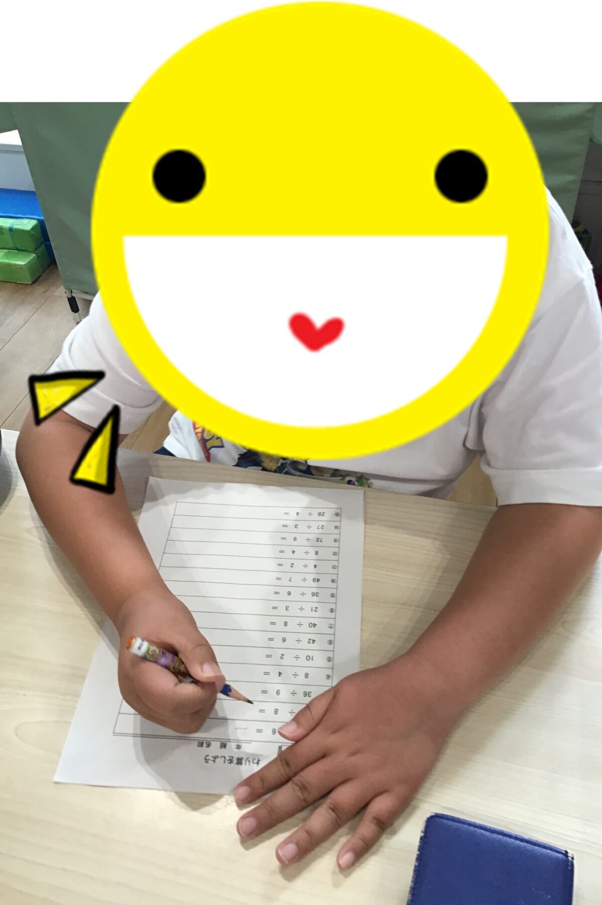 Child at a desk doing a math worksheet, pencil in hand; face covered by a large yellow smiley overlay.