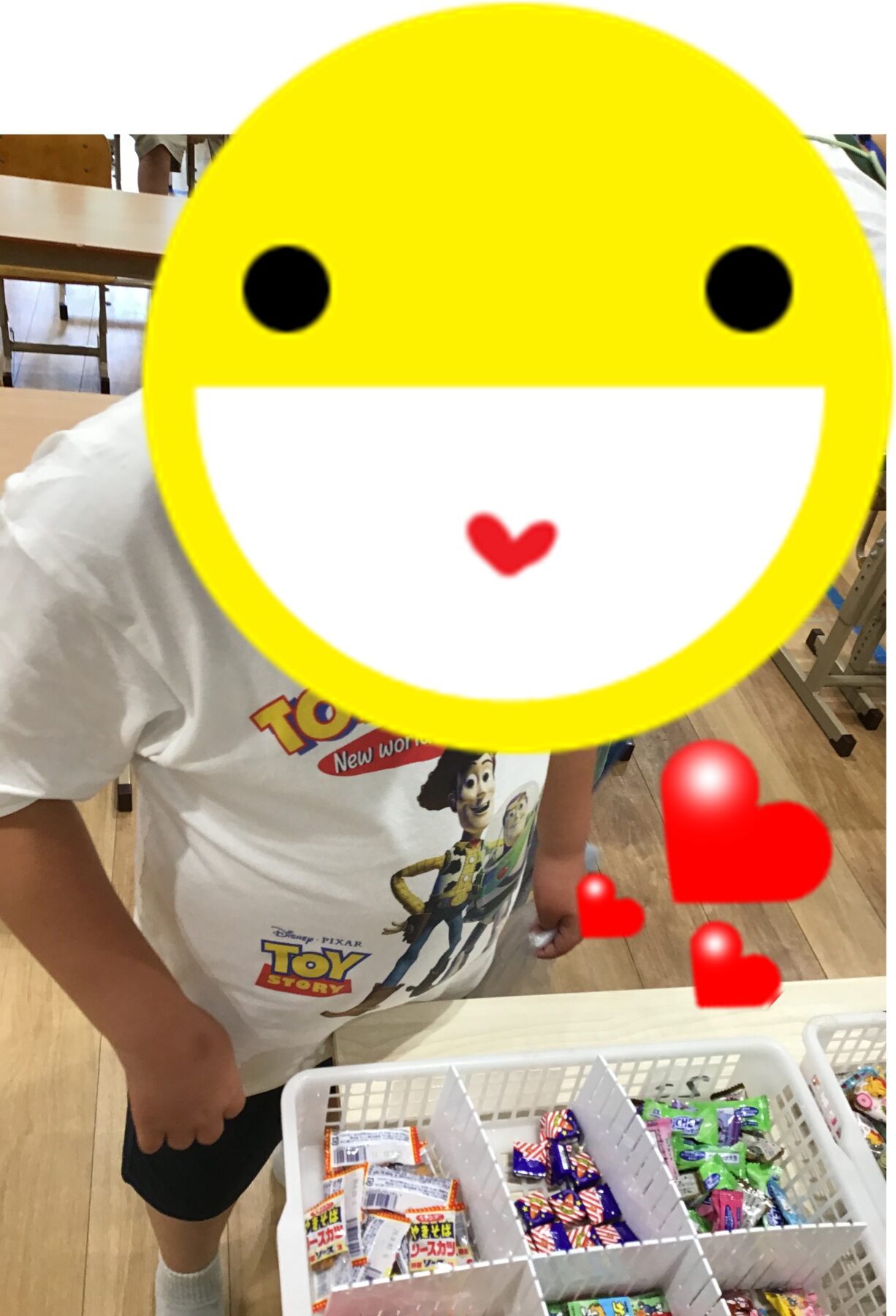 Child in a Toy Story shirt stands next to a candy display in a store; a large yellow smiling emoji overlay covers the face, with red heart stickers on the right side.