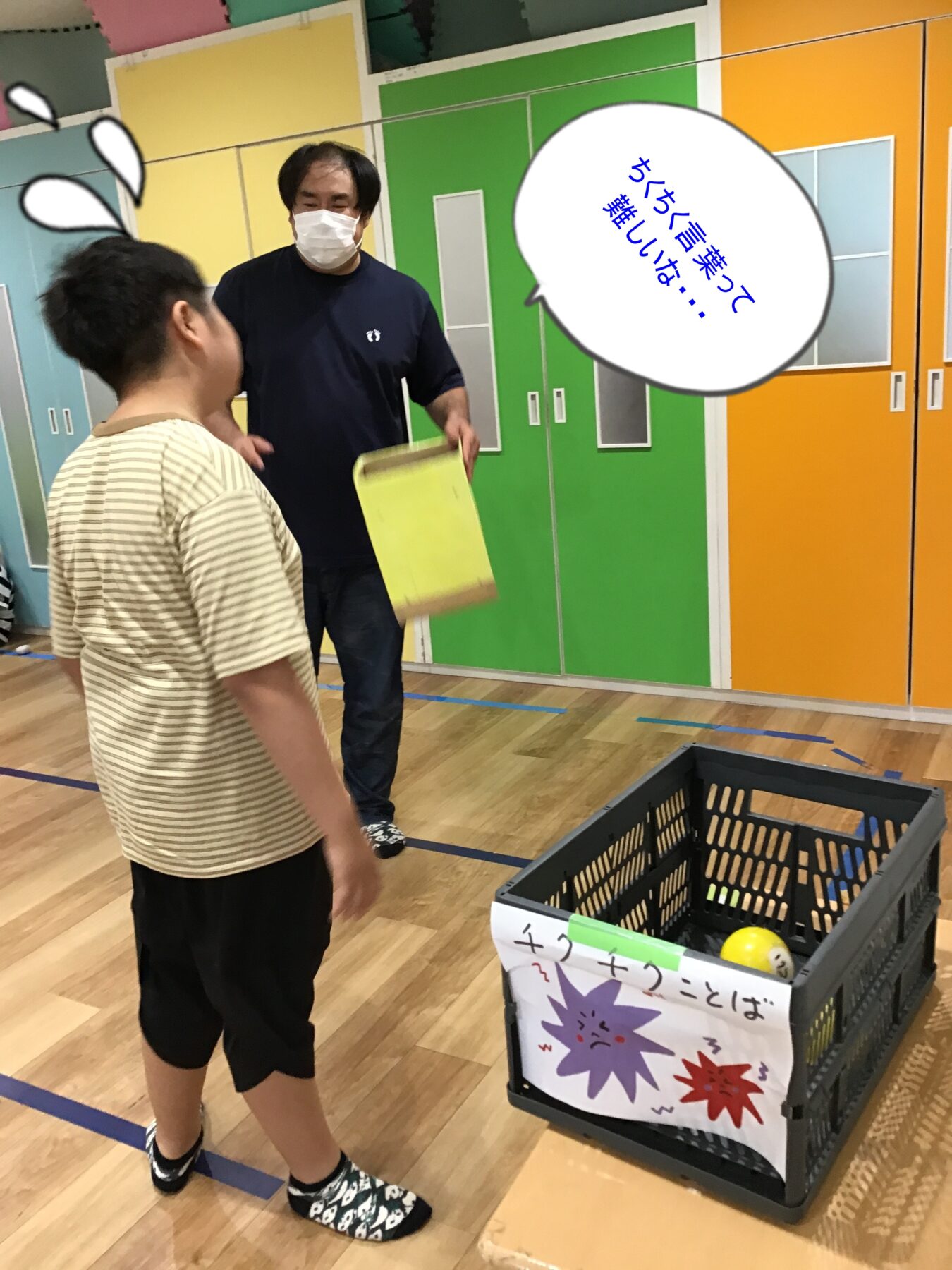 Two people in a colorful room: a man wearing a mask hands a yellow folder to a younger person in a striped shirt, near a crate with a sign in front.