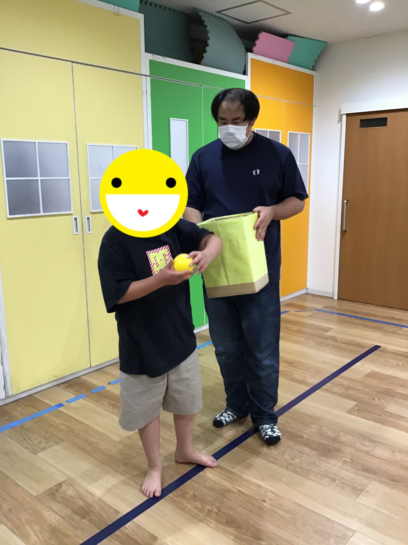 Adult in a dark shirt hands a yellow paper bag to a child; the child holds a small yellow ball in a room with bright yellow/green cabinets.