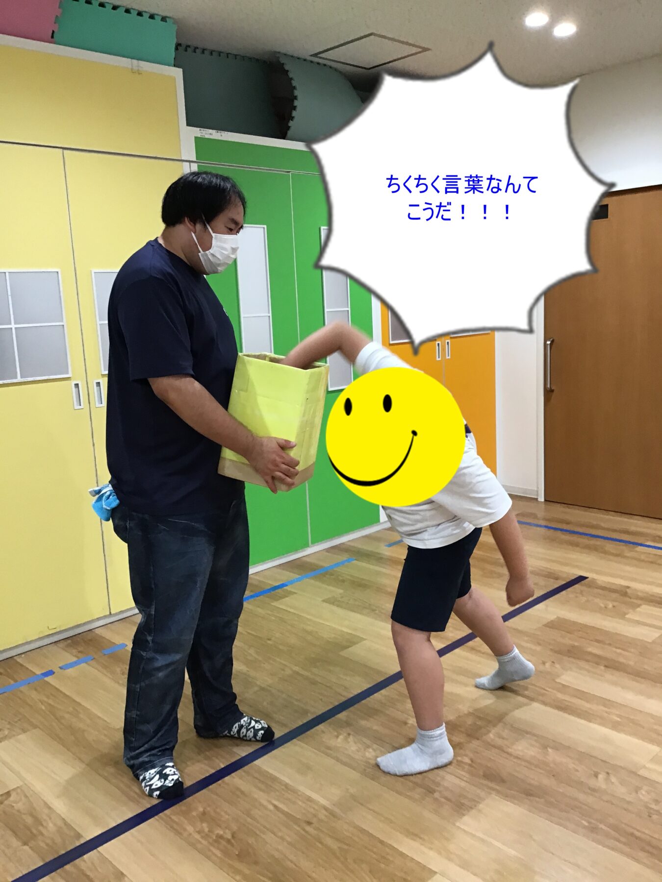Man wearing a mask hands a yellow box to a child in a gym; child leans forward toward the box with a yellow smiley face overlay, colorful lockers in the background.