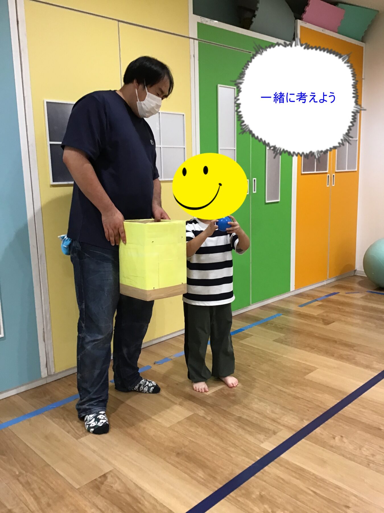Adult with mask helping a child in a colorful playroom; child holds a blue ball near a yellow box/container.