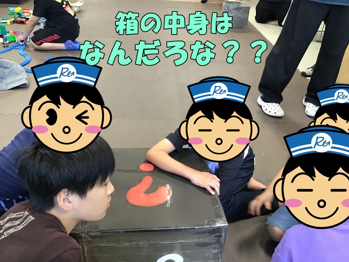 Children sit and kneel on a foam mat around a taped cardboard box, a red curved toy on top, with Japanese text asking what's inside.