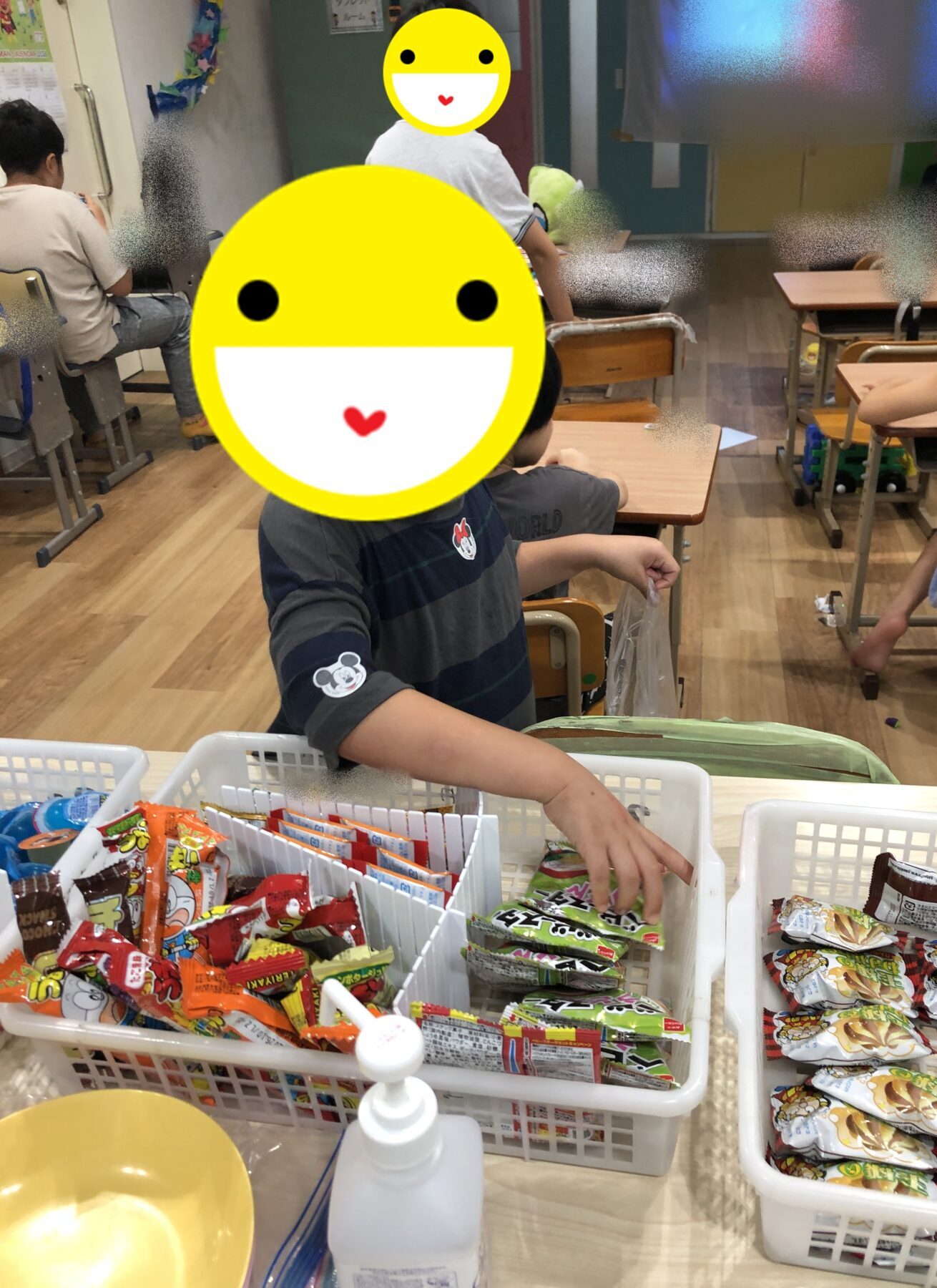 Classroom scene with children at desks; snacks in white baskets, faces obscured by large emoji stickers.