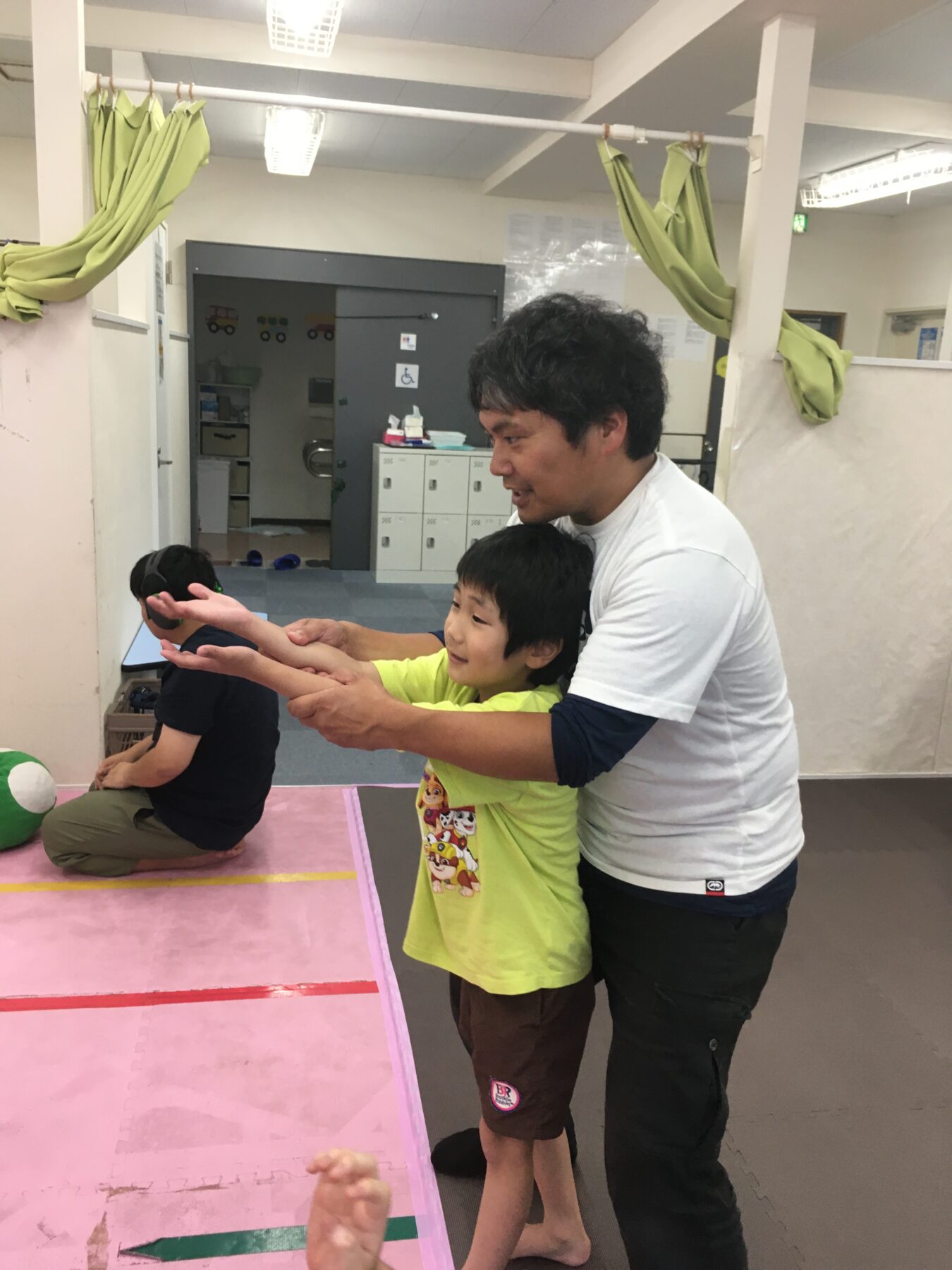 Adult man guiding a smiling boy as they extend their arms forward in a playroom/gym with pink mats and equipment in the background.