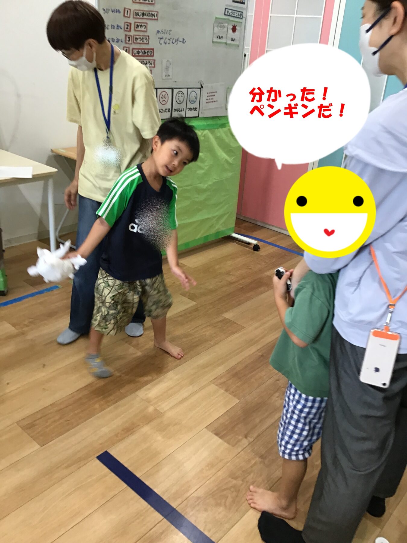 Young boy in a green and navy shirt throws a white stuffed toy as adults in masks look on in a bright classroom; speech bubble reads '分かった!ペンギンだ!' (I got it! It’s a penguin!).