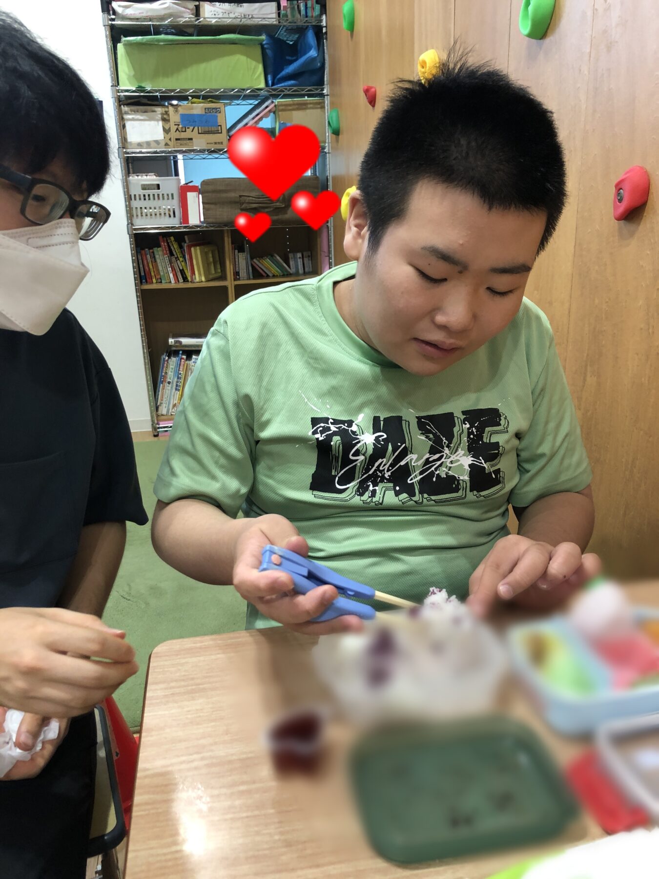 Boy in a light green shirt cutting with blue scissors at a table, another masked person beside him; shelves with boxes and books and a climbing wall in the background.