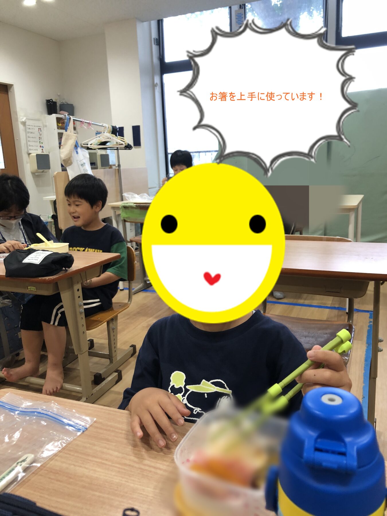 Classroom dining scene: a boy smiles while using chopsticks at a desk, with other kids eating nearby; a large yellow emoji covers a face and a speech bubble shows Japanese text 'お箸を上手に使っています!'