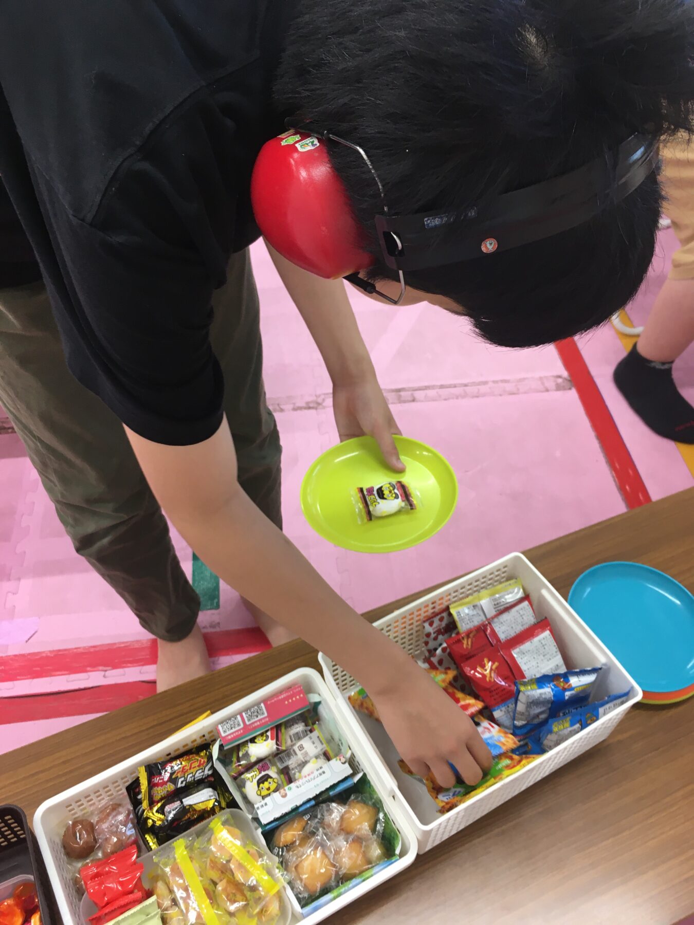 Person wearing red earmuffs and glasses bends over a table, selecting snack packs from white baskets on a pink floor mat.