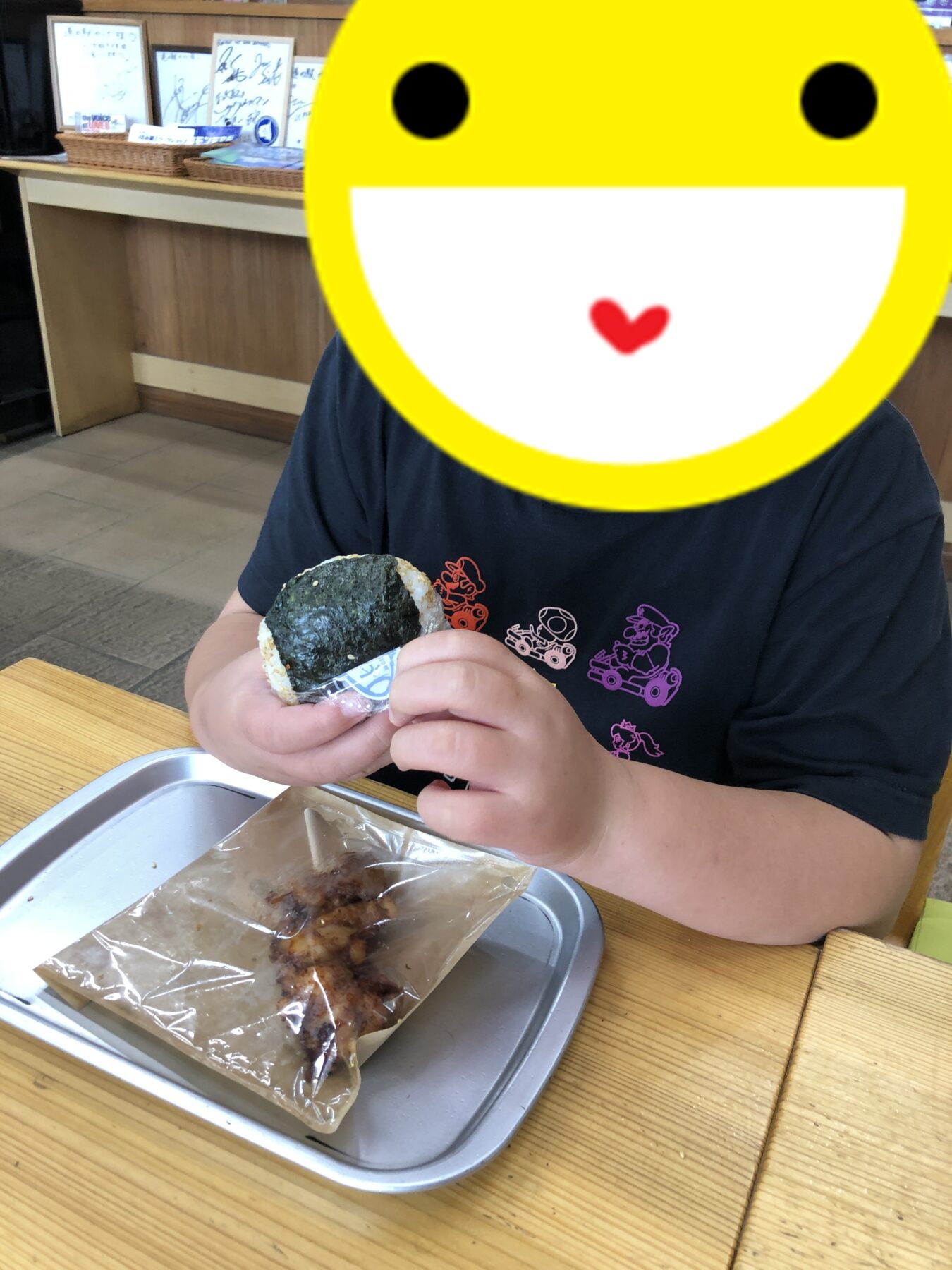 Person at a wooden-table restaurant holds a seaweed-onigiri (rice ball) in a wrapper, with a large yellow smiley image covering their face; a metal tray with plastic-wrapped food sits in front.