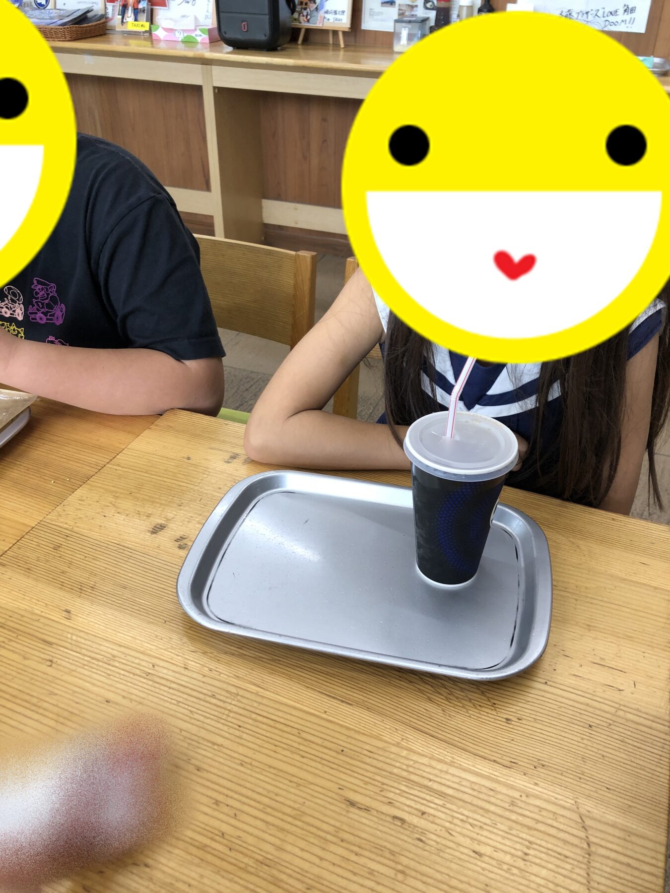 Two people sit at a wooden table in a casual dining area; a blue beverage cup with straw rests on a metal tray in front of them, their faces covered by yellow smiley overlays.