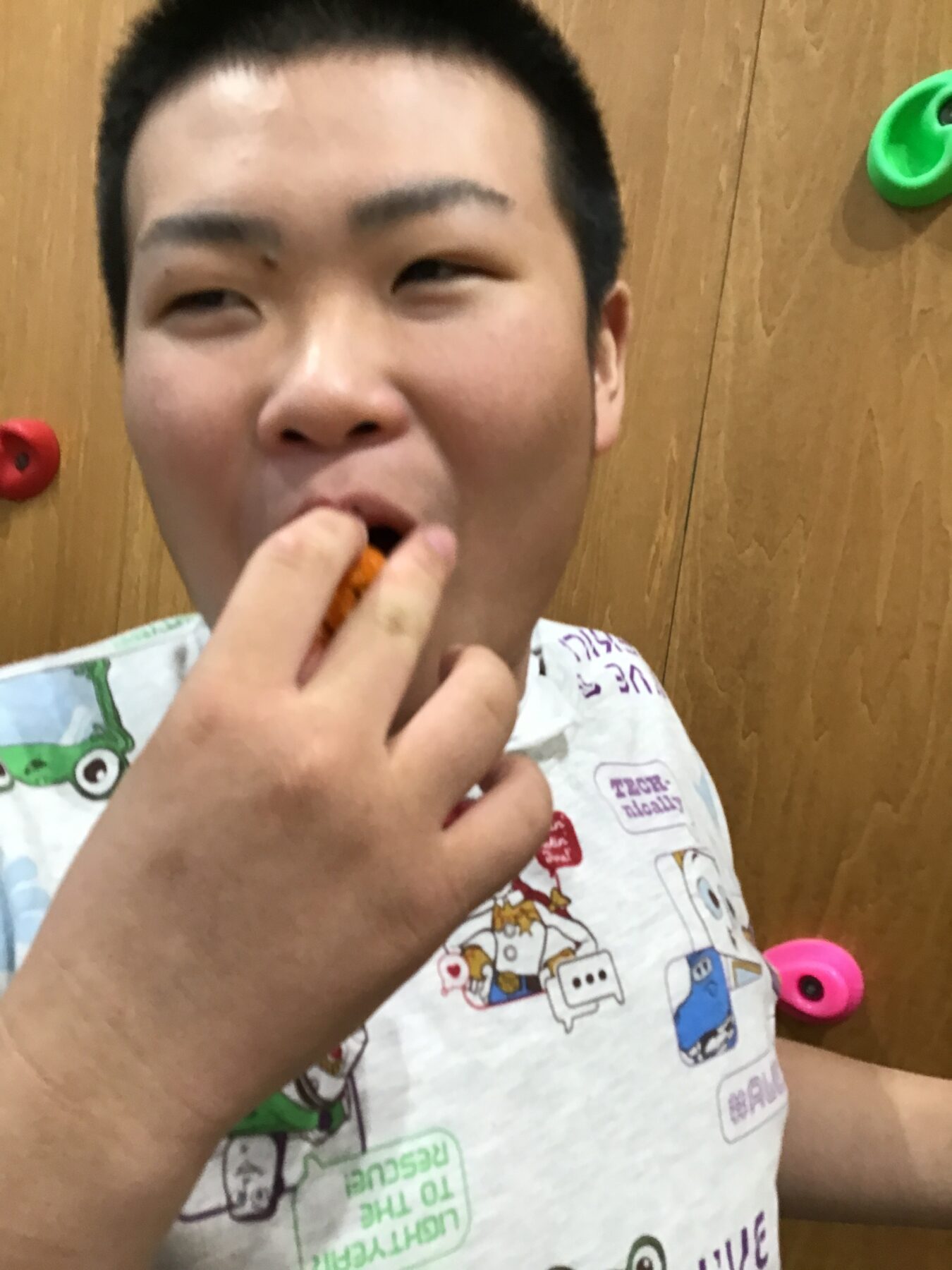Close-up of a boy biting an orange snack, looking at camera with a wooden wall behind him.
