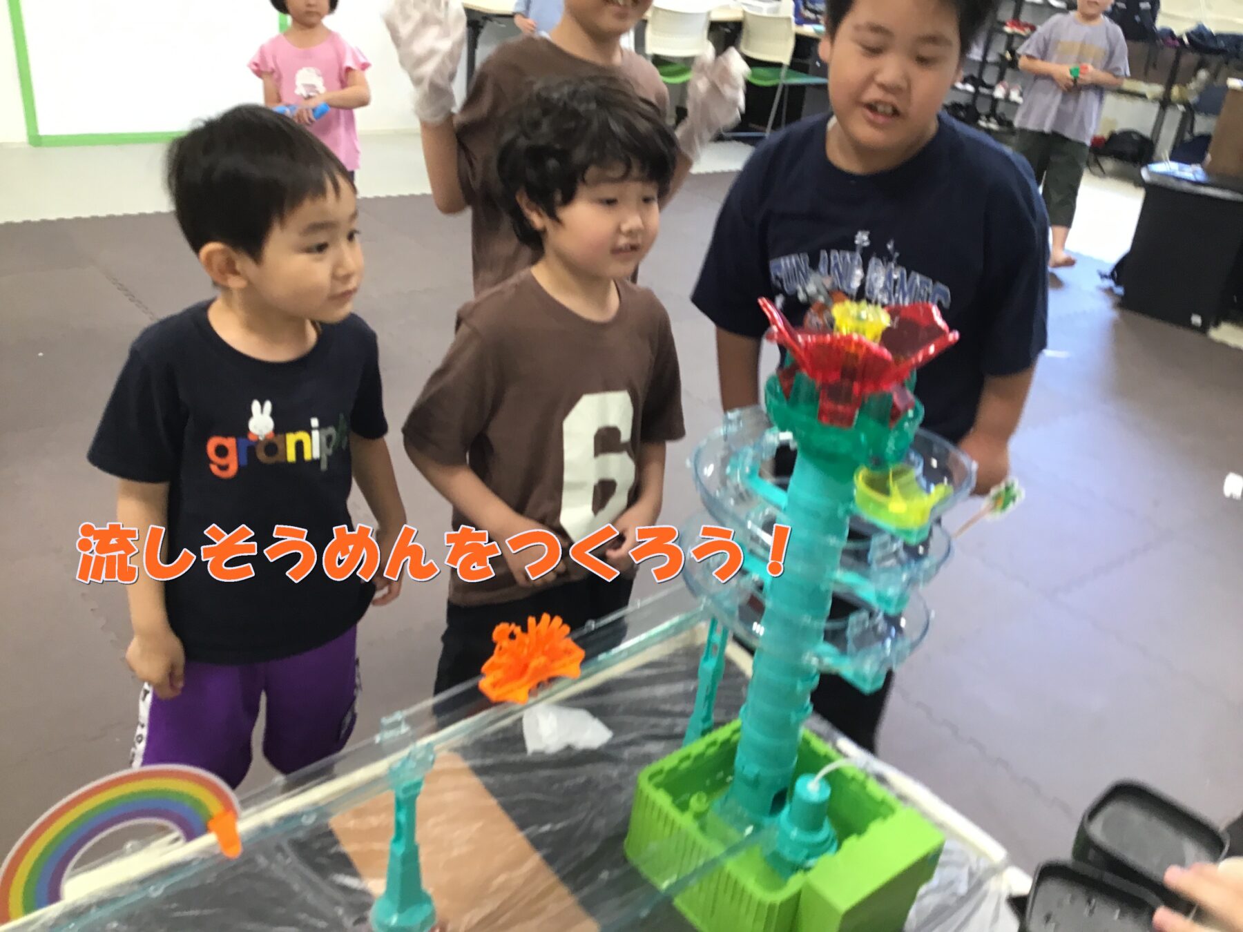 Three children watch a colorful DIY water-flow sculpture during a nagashi somen activity in a classroom setting