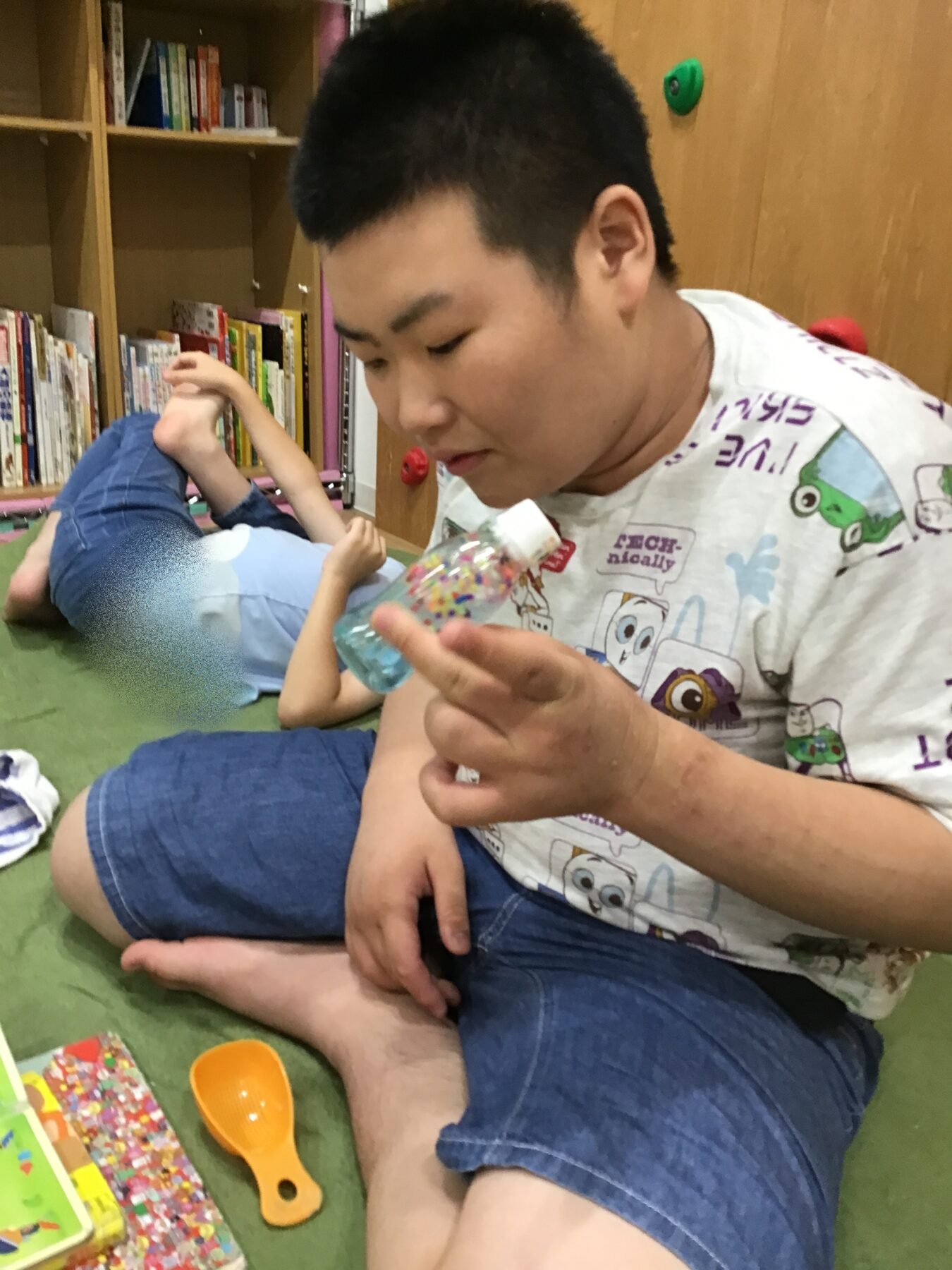 Young man in a printed T‑shirt sits on a green carpet, holding a clear bottle with colorful decorations inside while another child lies on the floor in the background among bookshelves and toys.