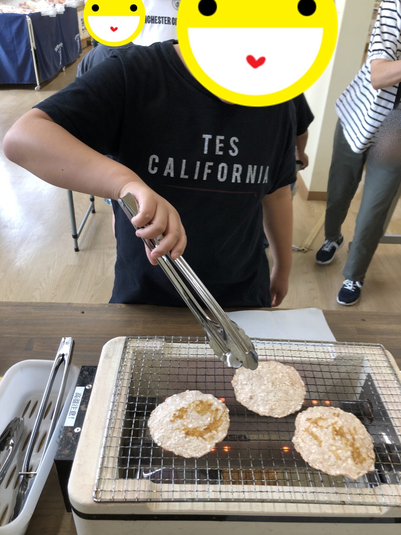 Person using tongs to grill breaded patties on a countertop grill; wearing a dark T-shirt with 'TES CALIFORNIA' text, emoji faces cover their head for privacy.