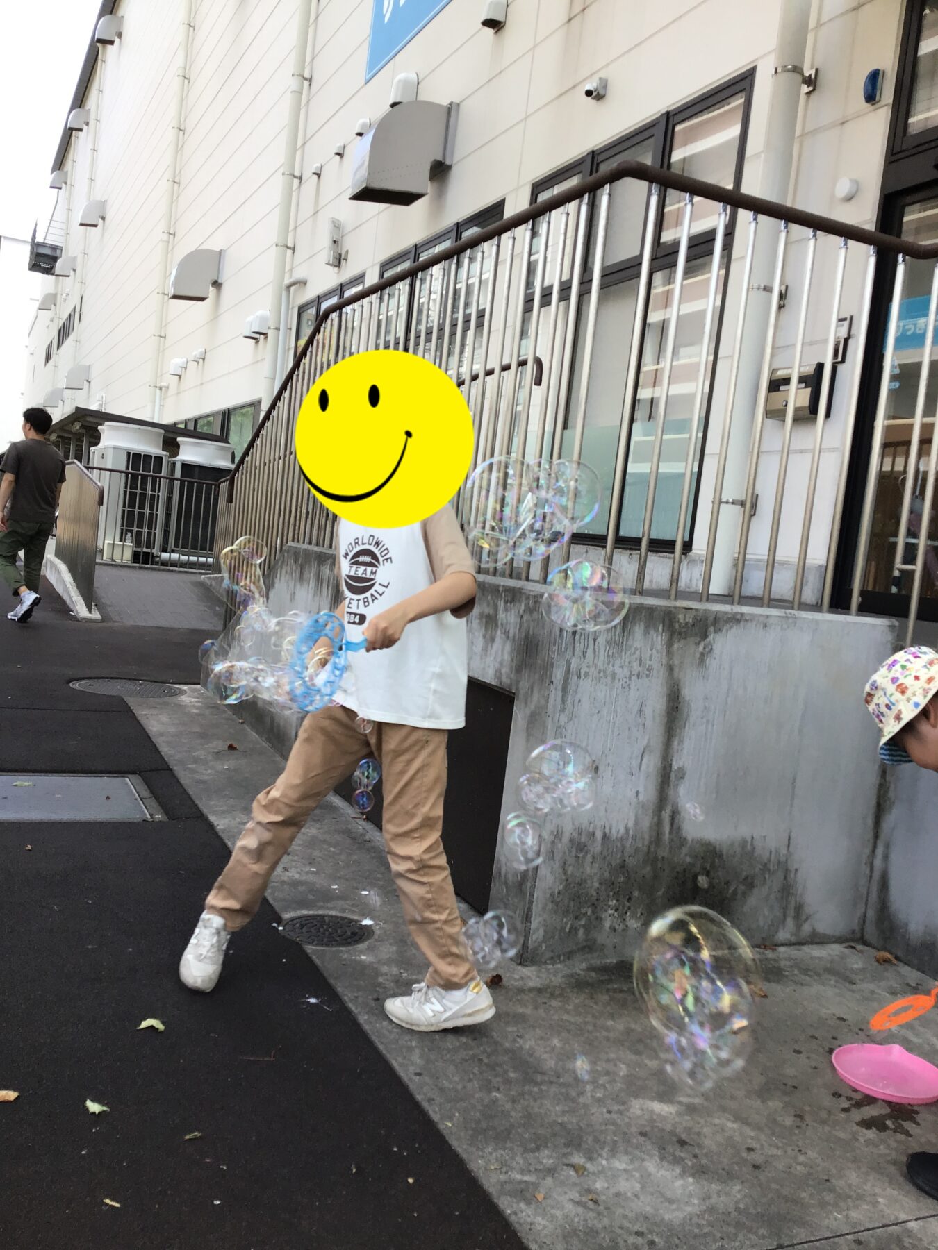 Child blowing bubbles on a sidewalk beside a modern building with a metal railing outside an entrance.