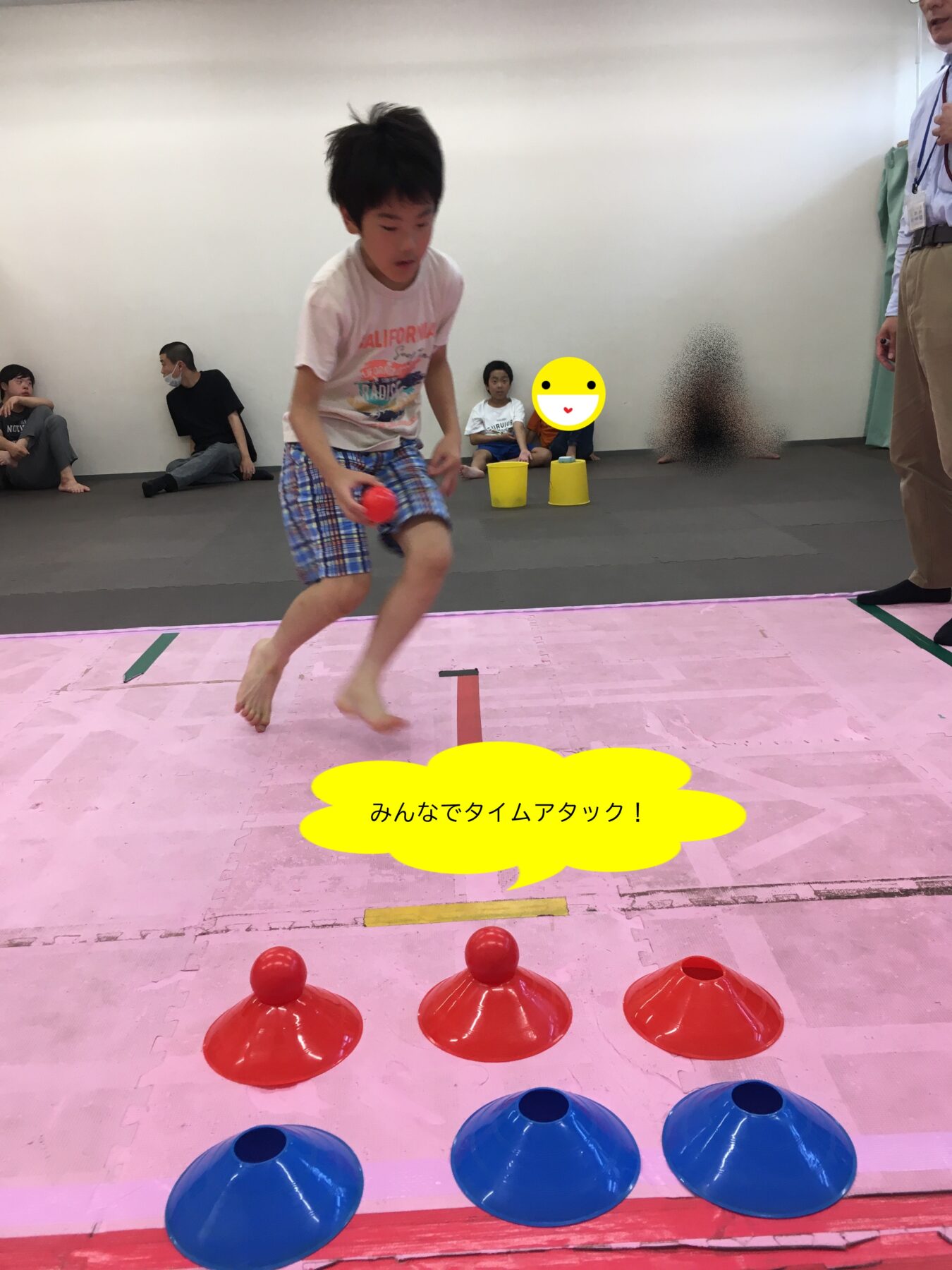 Boy in shorts jumping on a pink gym mat, holding a red ball, with red and blue cones lined up and spectators watching in the background.