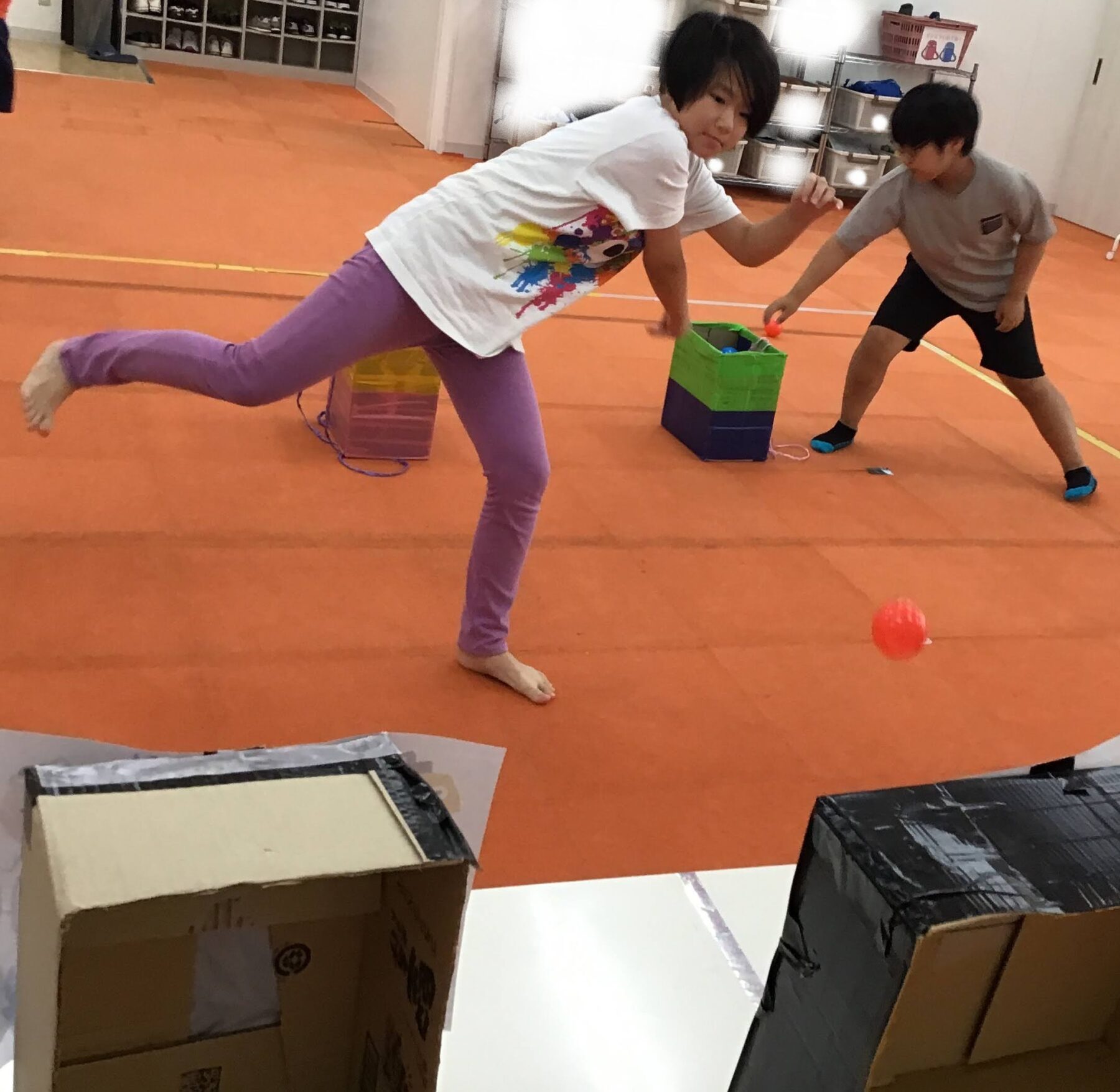 Two kids play in a gym-like room with cardboard boxes; a girl in a white shirt and purple pants balances on one leg while reaching toward a small orange ball, a boy in gray and black shorts extends a hand nearby.